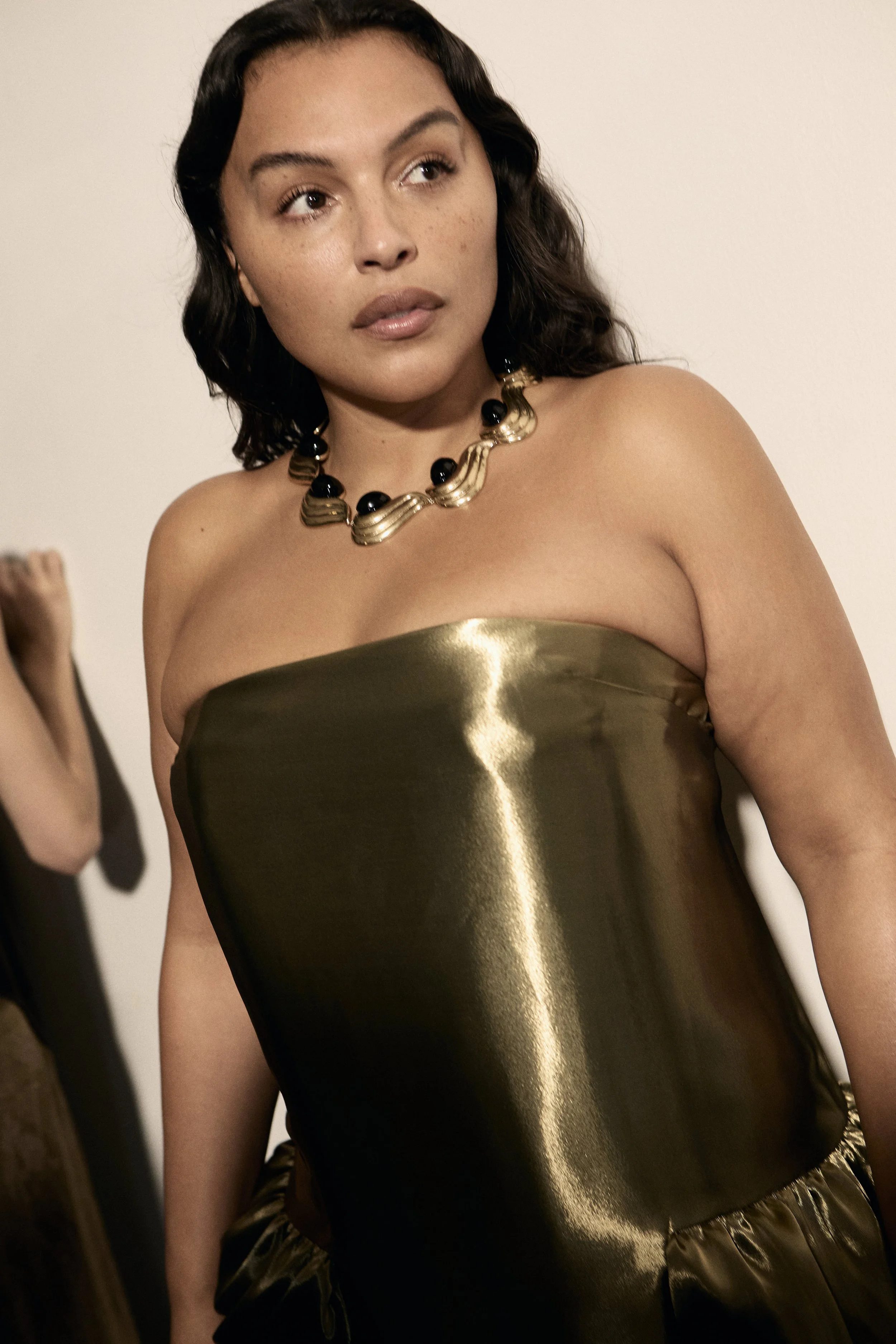A woman with dark wavy hair and freckles wearing a strapless shiny gold dress and a chunky black and gold necklace.
