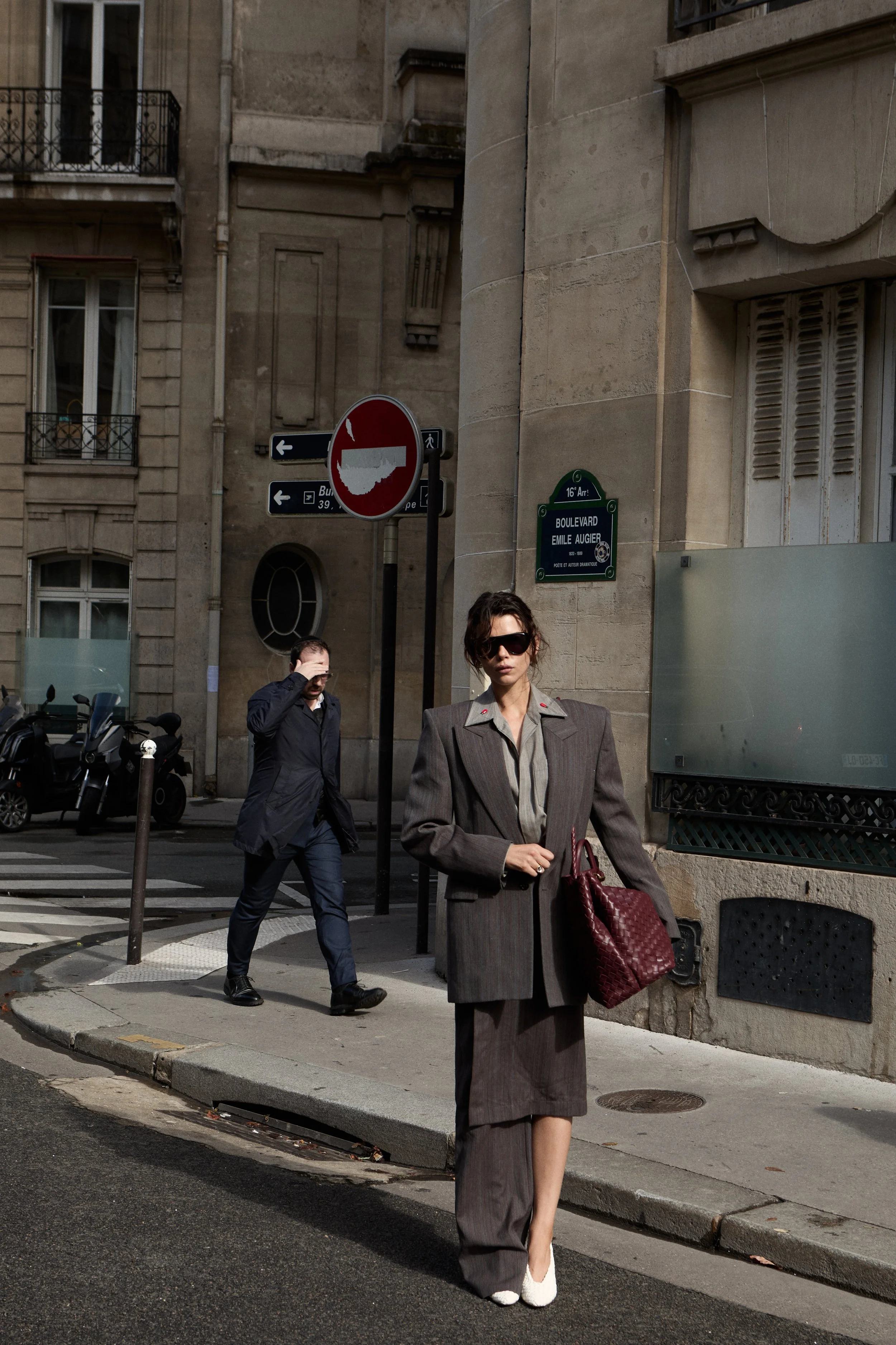 Model Georgia Fowler in a gray business suit carrying a red handbag walks on a city street with a man in a dark jacket walking behind her. A street sign indicates the location as Boulevard Emile Augier on 16th Arrondissement in Paris.