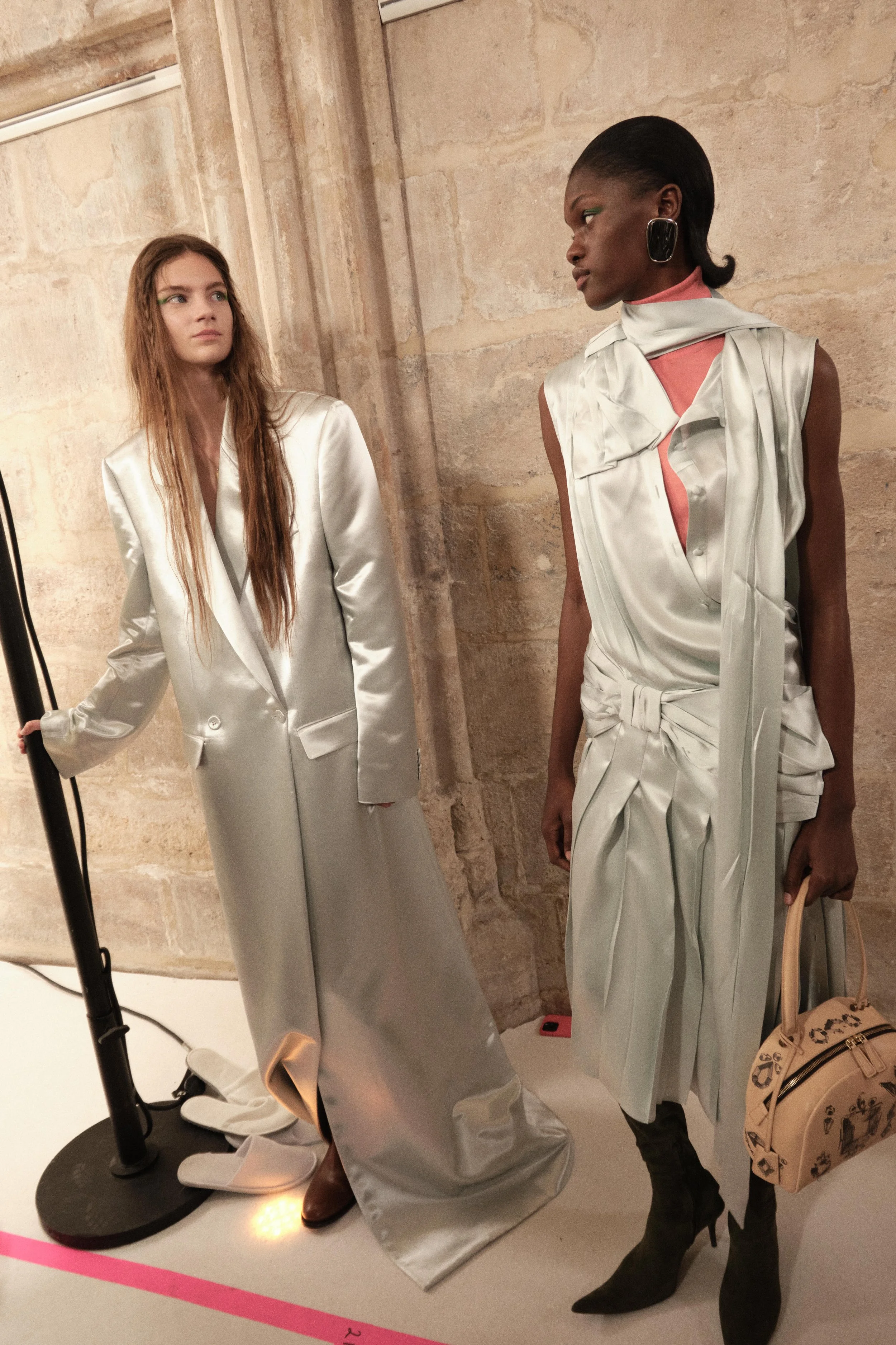 Two models in shiny, silver designer outfits at a fashion show backstage. One has long, wavy hair and is wearing a long blazer dress, while the other has short hair, large earrings, and is holding a handbag.