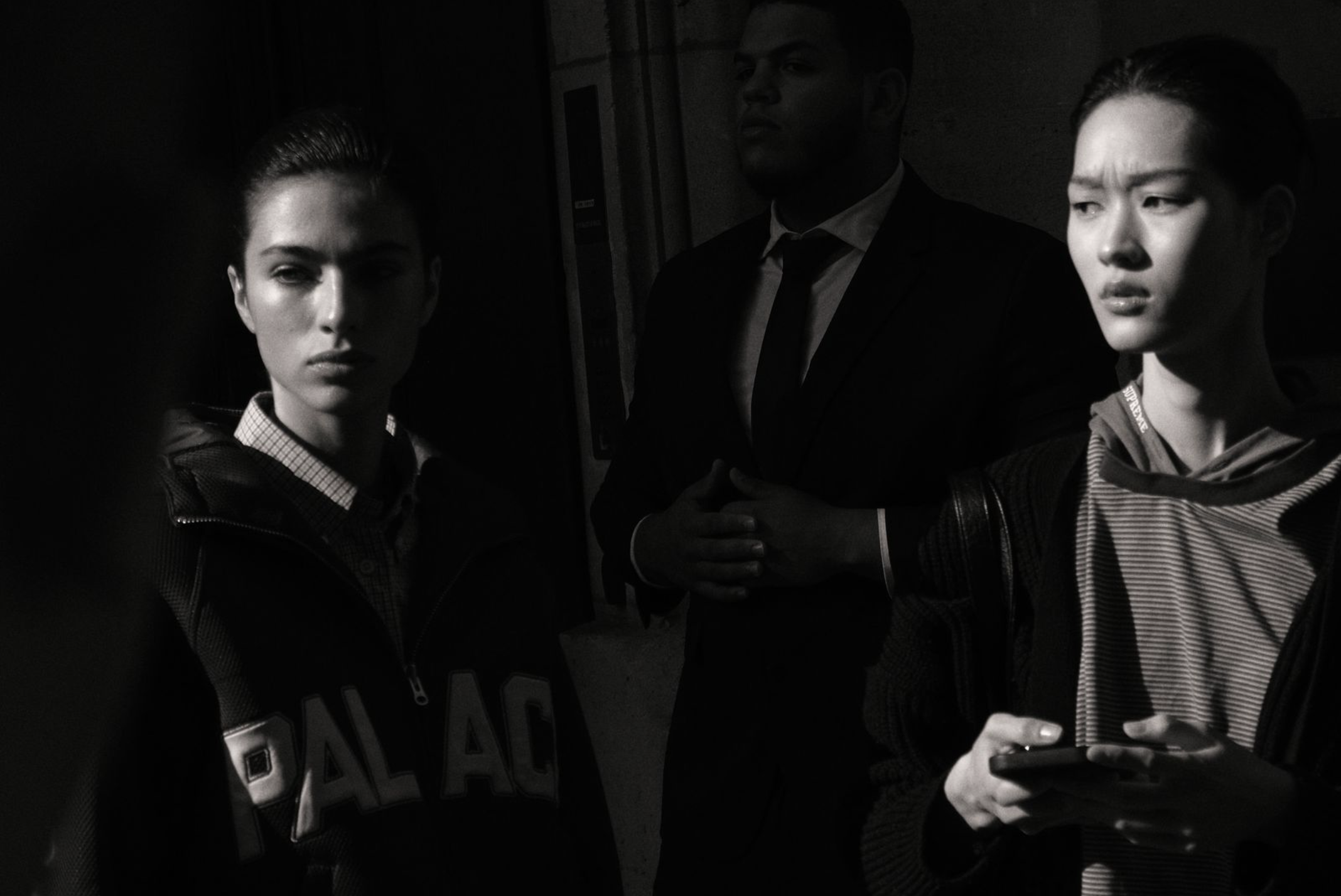 Three young adults appear in a dark setting with low lighting. Two women are in the foreground, one on the left with short hair and a hoodie, and one on the right with short hair and a striped hoodie, holding a phone. A man is in the background, wearing a suit and tie, with hands clasped. The lighting adds dramatic shadows, and the mood is serious or contemplative.