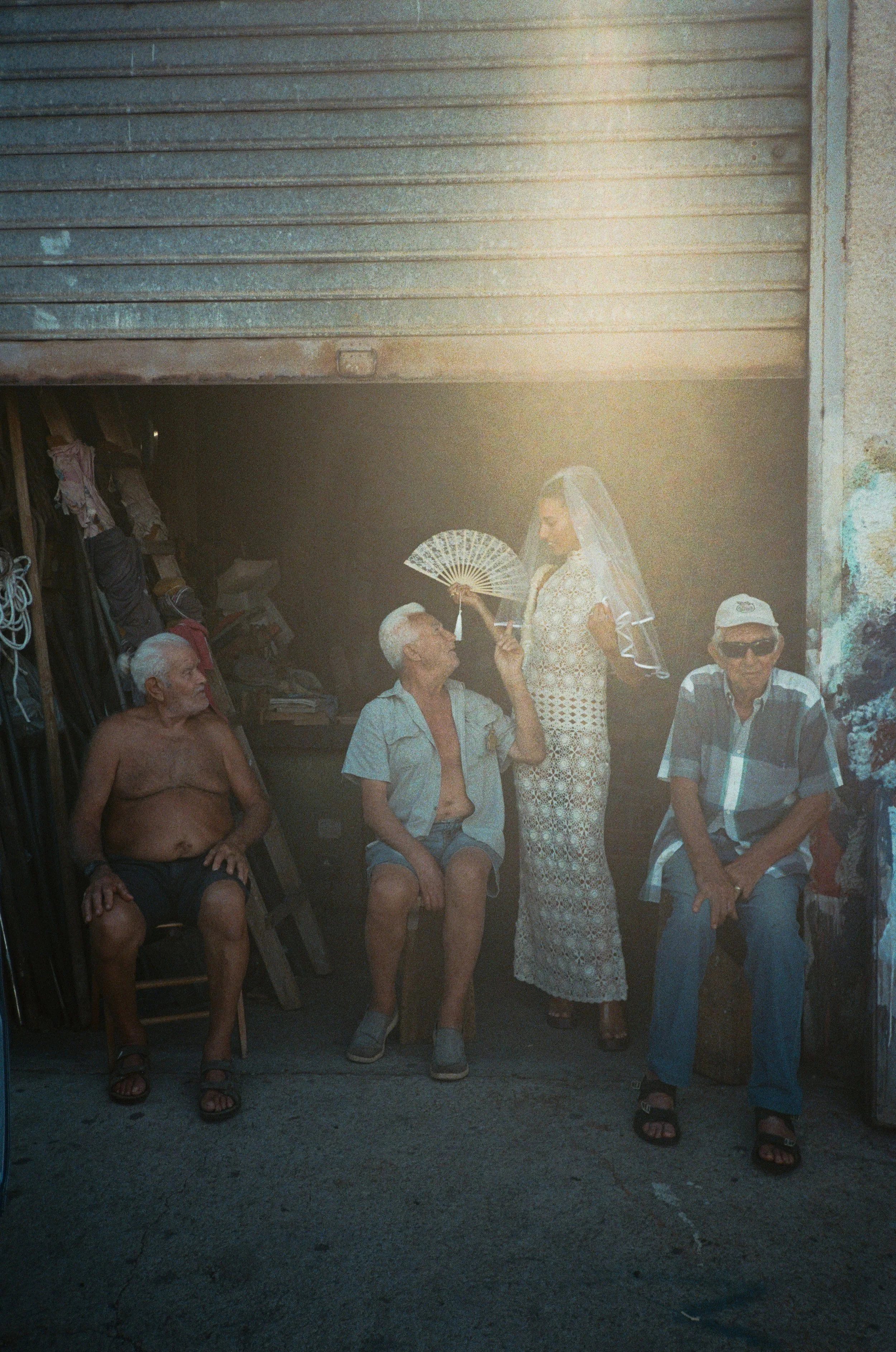 A woman in a floral dress and veil stands in front of three elderly men sitting on chairs in a garage. She is holding a fan and looking at one man. The men are casually dressed, with one shirtless. The garage has various items stacked inside, and sunlight filters into the scene.