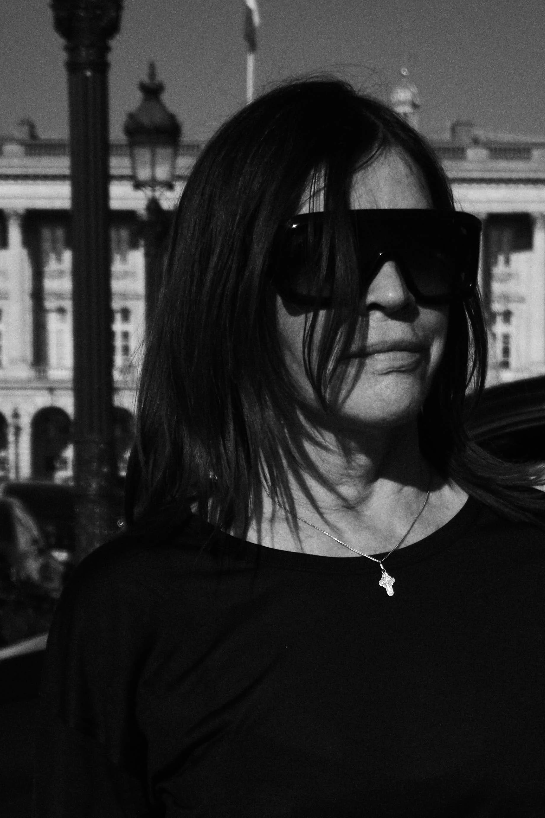 A black and white photo of a woman wearing large sunglasses and a necklace with a cross pendant, standing outdoors with a building and lampposts in the background.