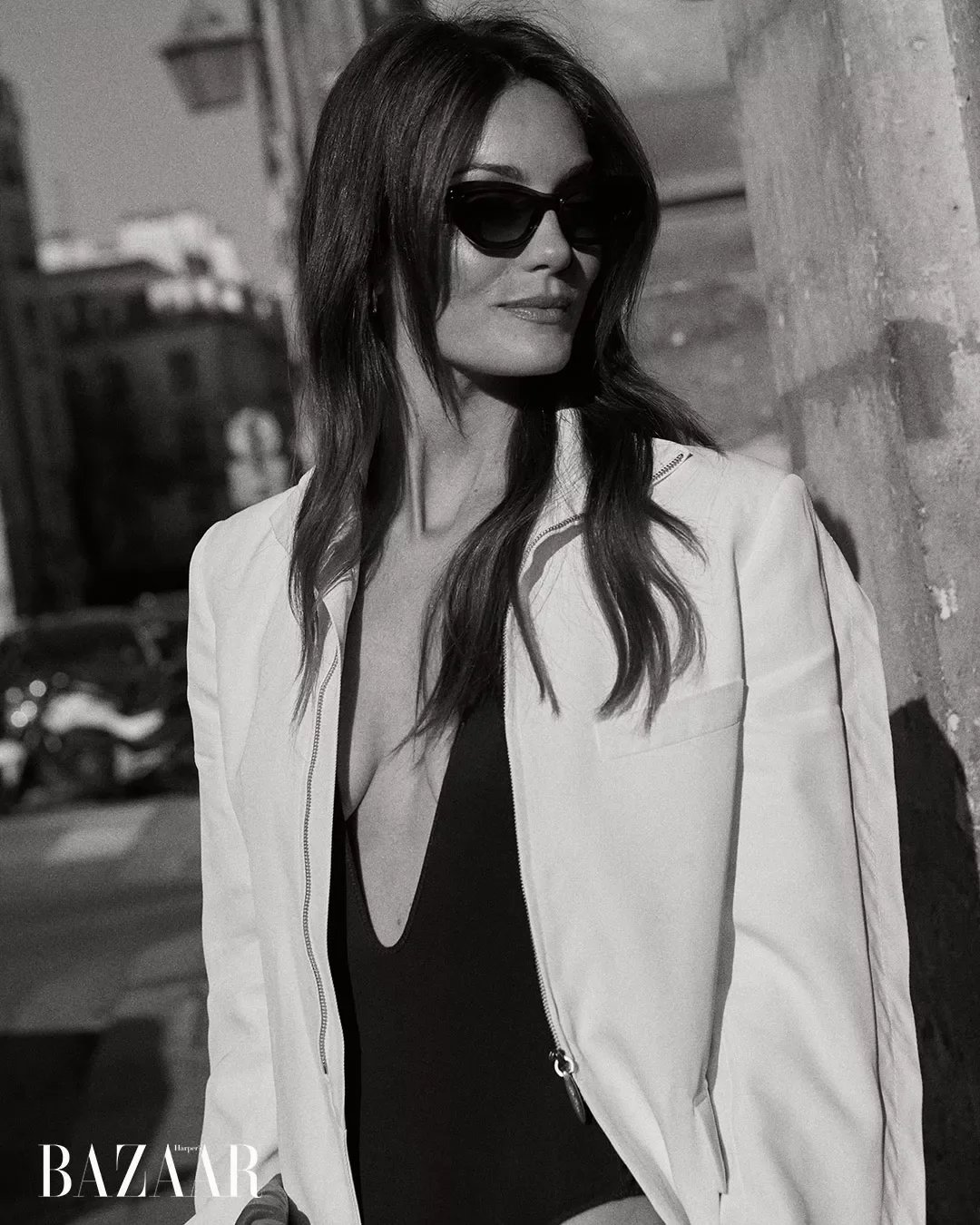 A black and white photo of a woman wearing sunglasses, a white jacket, and a deep V-neck top, leaning against a wall outdoors.