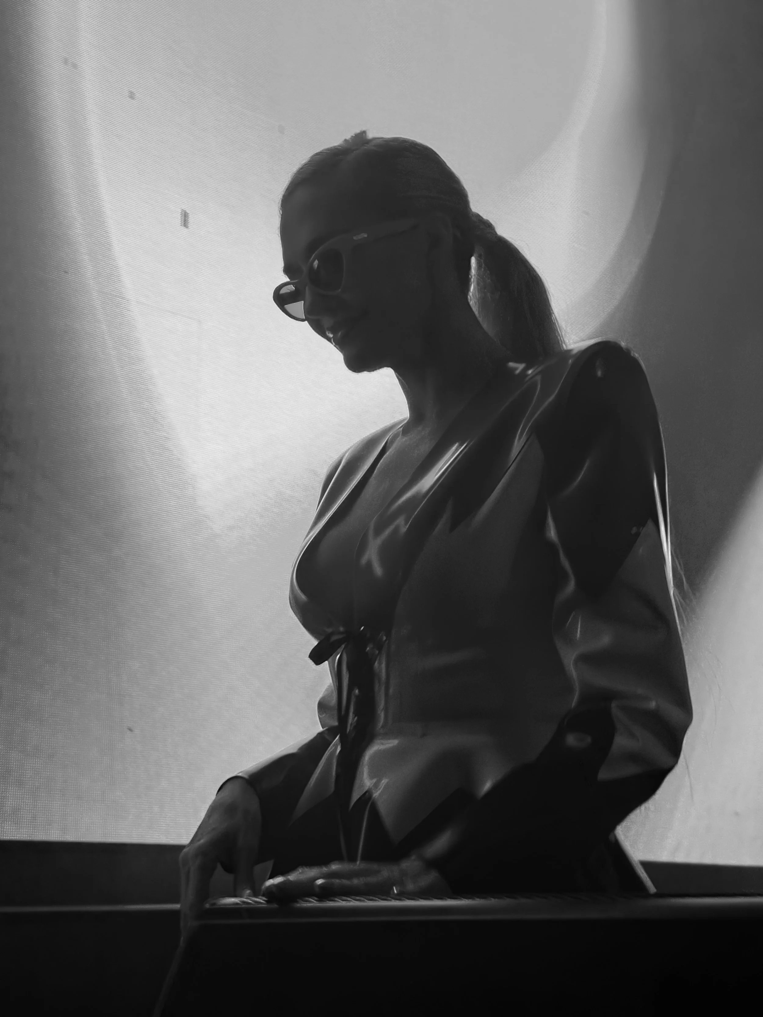 American singer and TV personality Erika Jayne wearing glasses and a shiny, possibly leather or latex, jacket and a ponytail is smiling while leaning on a surface, dj-ing.