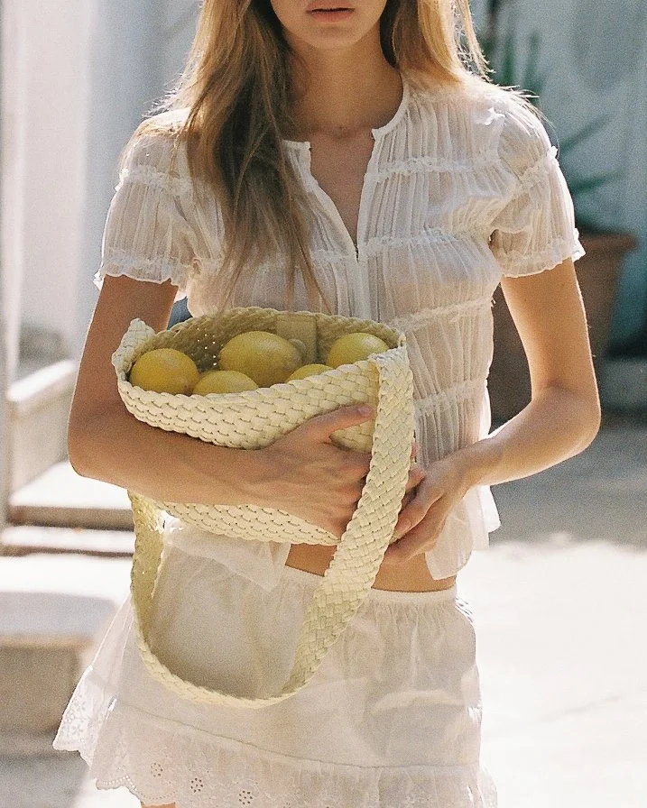 A woman holding a woven basket filled with lemons in a bright outdoor setting.