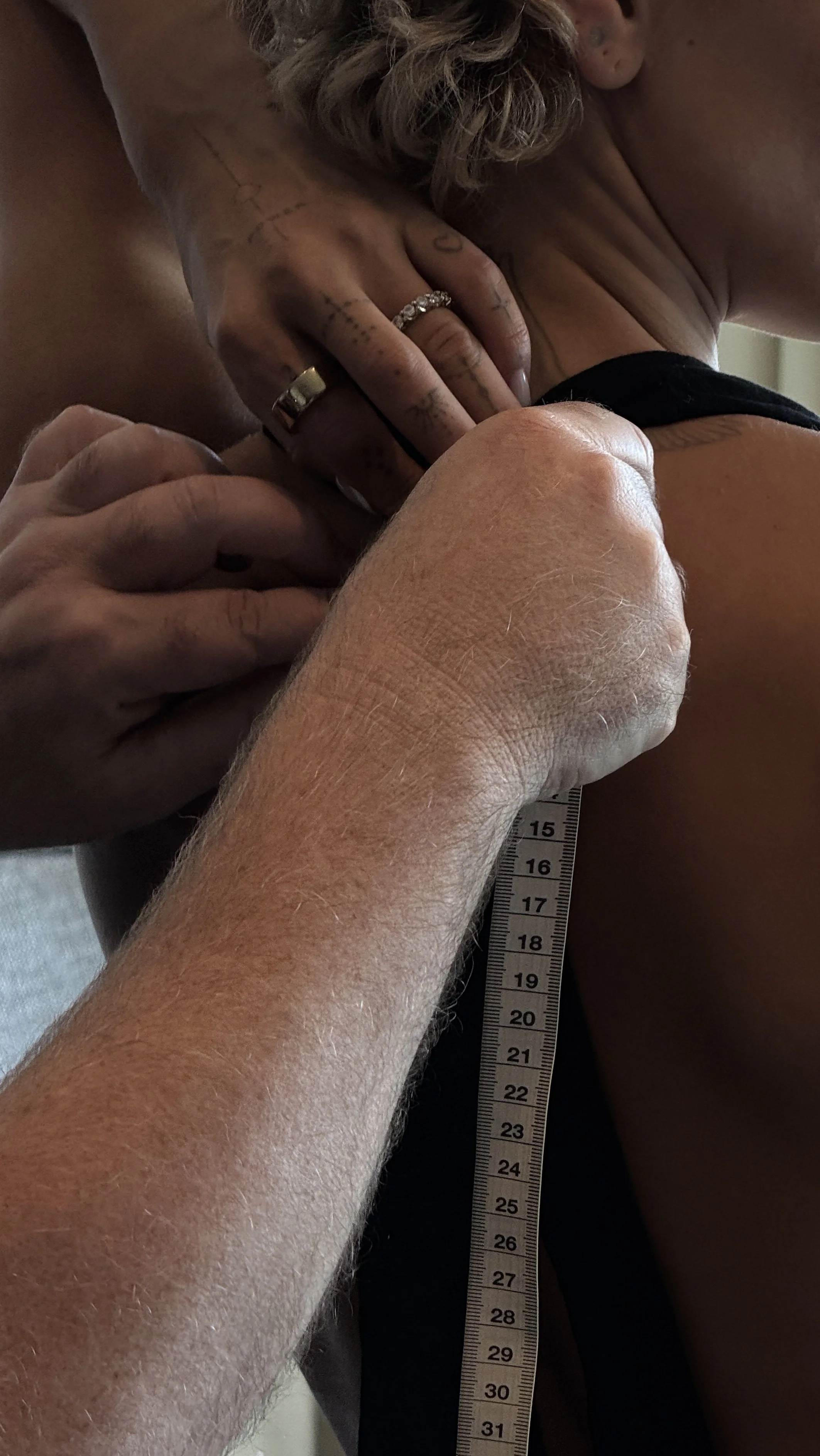 Close-up of a person measuring another person's shoulder with a ruler, with their hand resting on the person's shoulder, both appearing to be in an intimate setting.