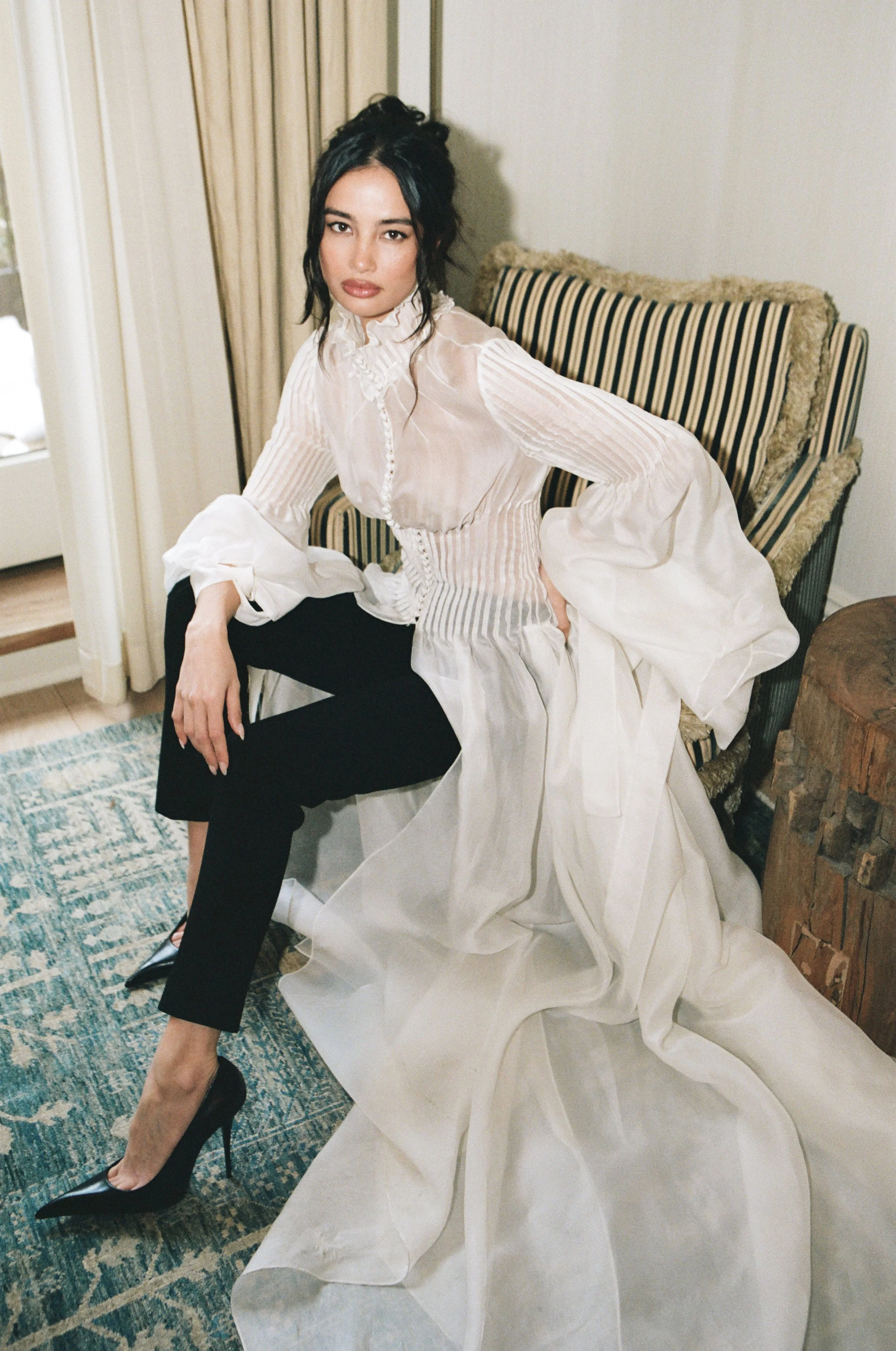 A woman sitting on a striped, upholstered armchair with ornate trim, wearing a sheer, white, ruffled blouse, black pants, and black high heels, with dark hair styled in loose waves, in a room with light-colored curtains and a side table.