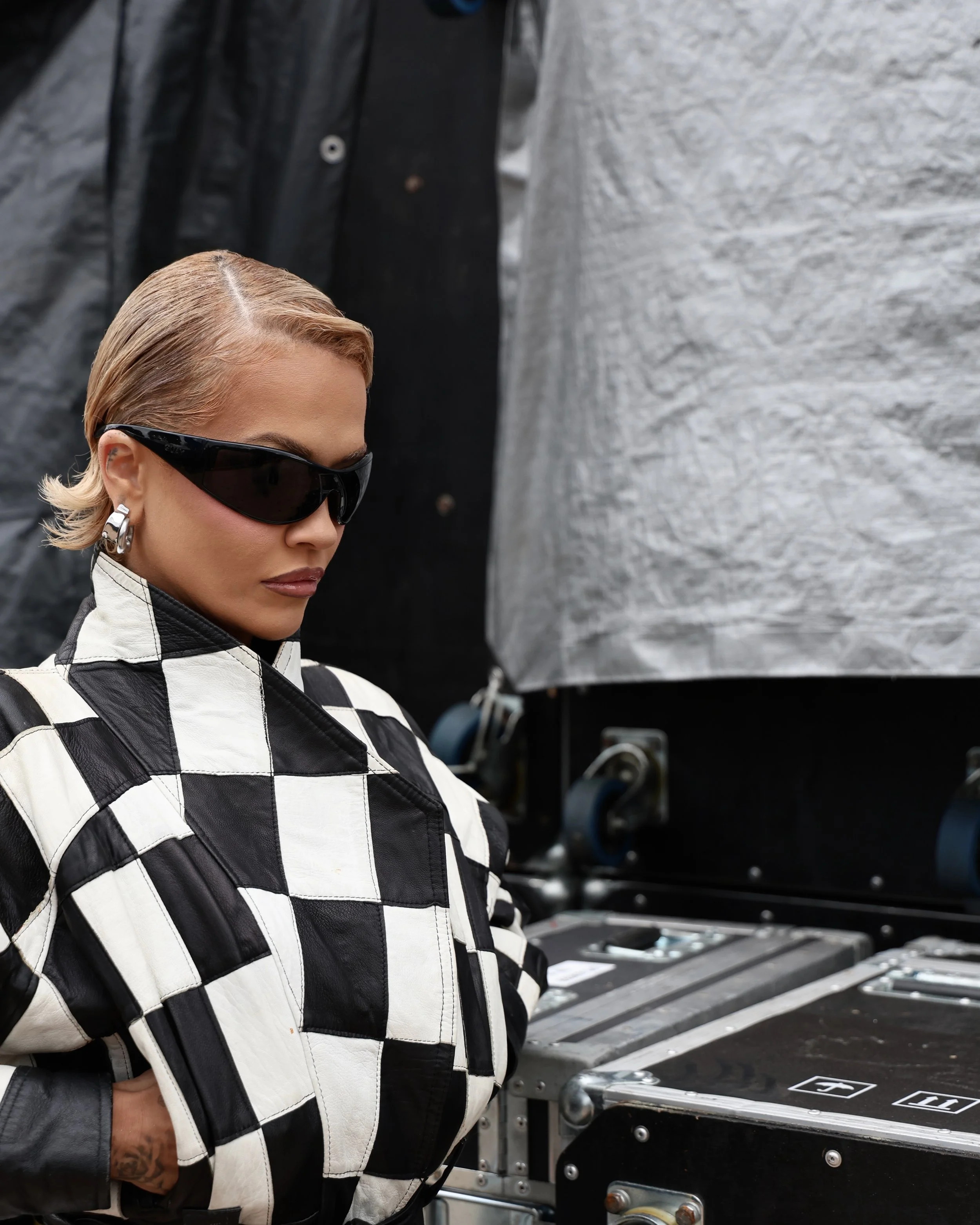 Singer Rita Ora at the Formula 1 Grand Prix 2026, wearing black sunglasses, gold hoop earrings, and a black and white checkered leather jacket.