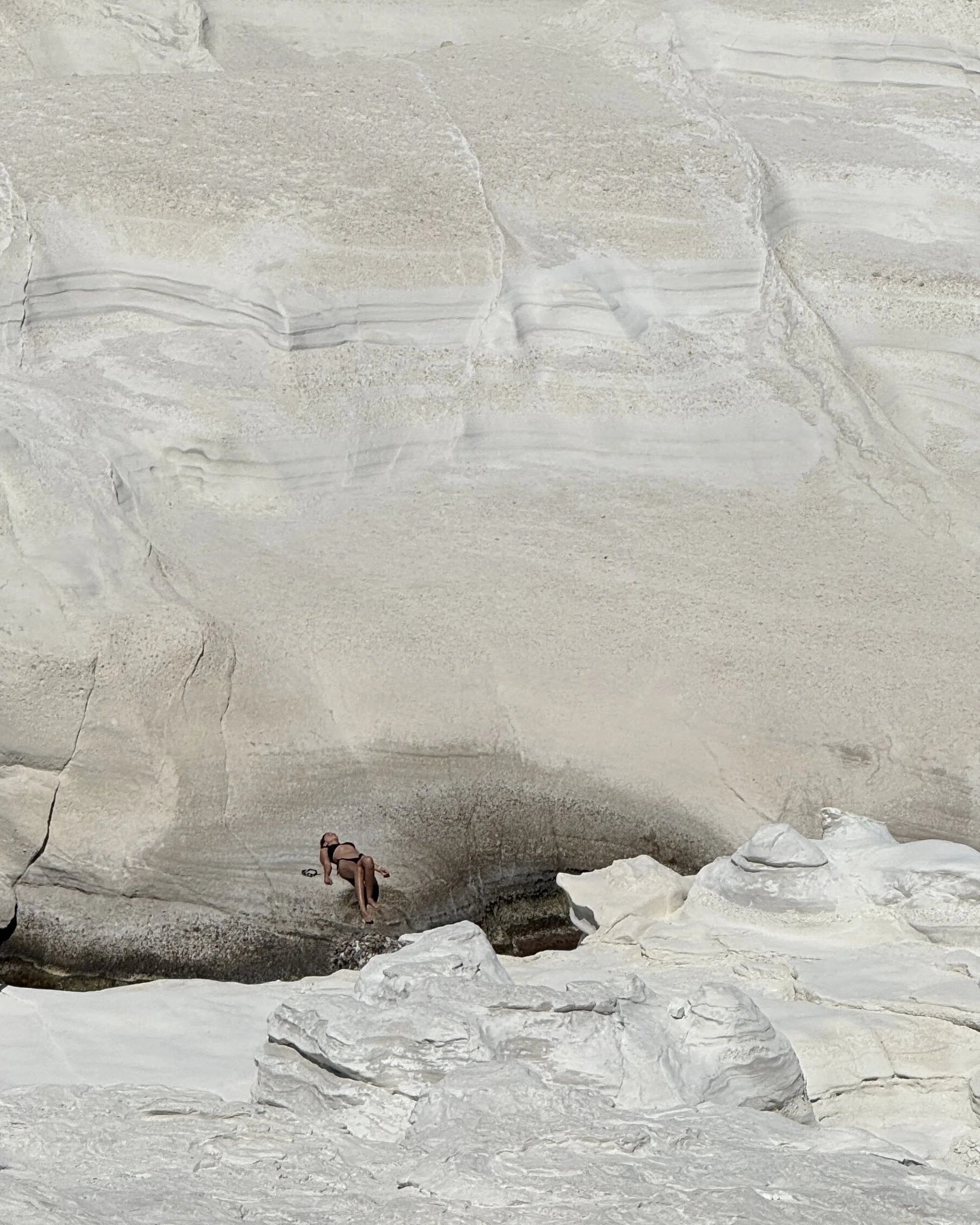 Person lying down on a large, smooth, light-colored rock formation in Milos, Greece, with a vast, beige sandstone wall in the background.