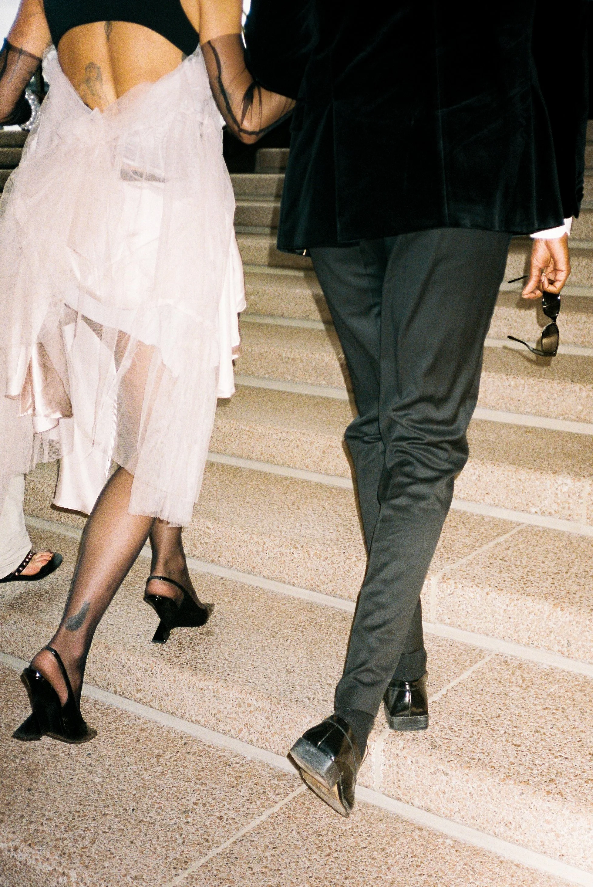A woman dressed in a sheer, white, layered dress with black heels and tattoos on her legs walking up stairs. A man next to her wearing black dress shoes, dark pants, and a black blazer, holding sunglasses.