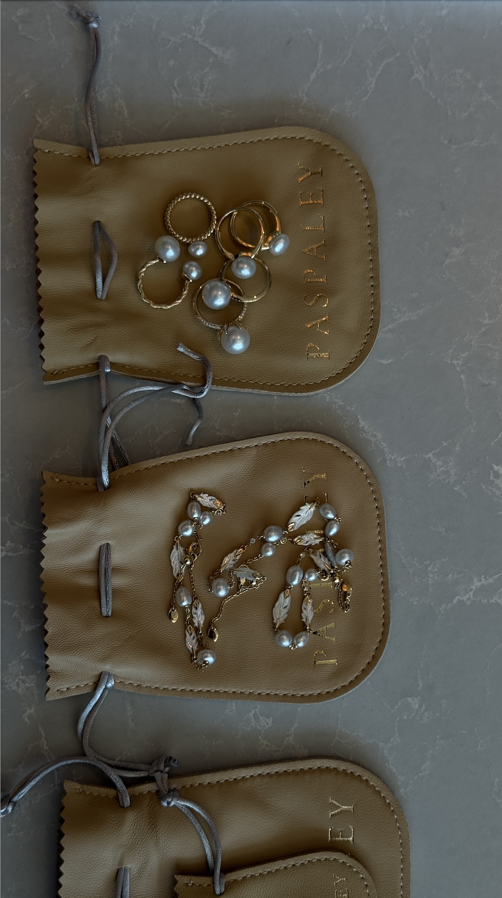 Three beige jewelry pouches with silver drawstrings, each containing jewelry from Paspaley Pearls. The top pouch has gold rings with pearl accents; the middle pouch has gold earrings with pearl and feather details.