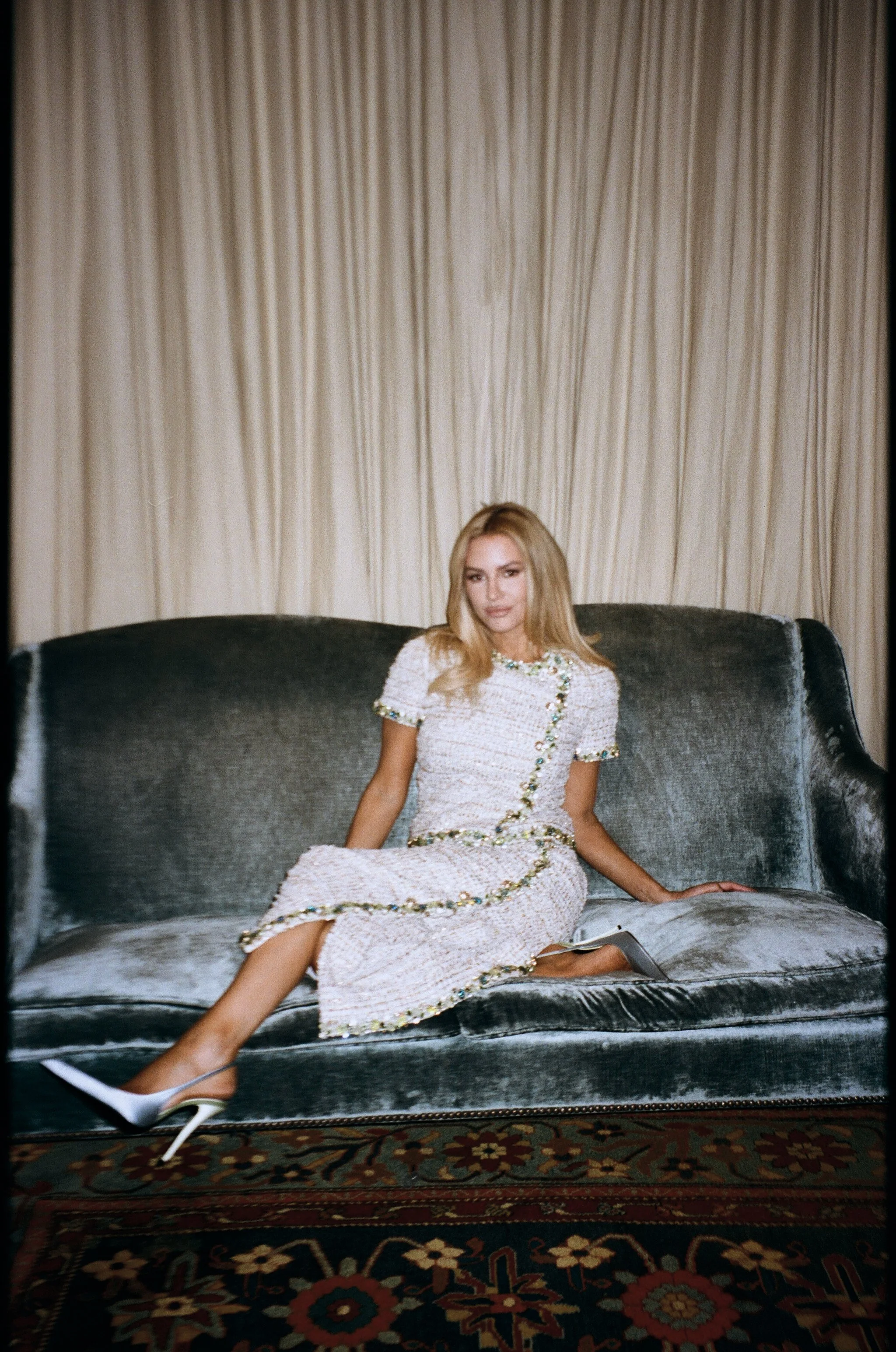 A woman in a white dress with embellishments sits on a vintage dark gray velvet sofa in front of beige curtains.