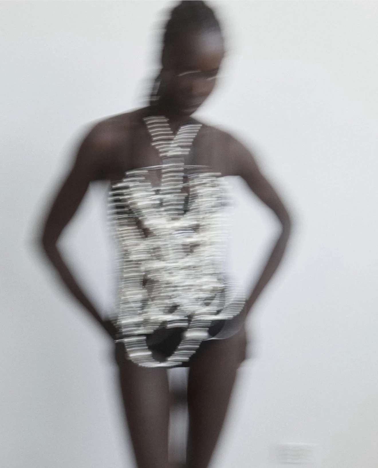 Blurry image of a person with dark skin wearing a strapless, silver, and black patterned dress and a large beaded necklace, standing in front of a plain light-colored wall.