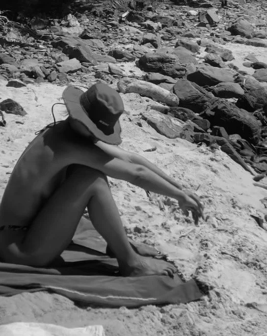 A person sitting nude on a towel at the beach in Byron Bay, wearing a hat, with rocky shoreline in the background. Film photography.
