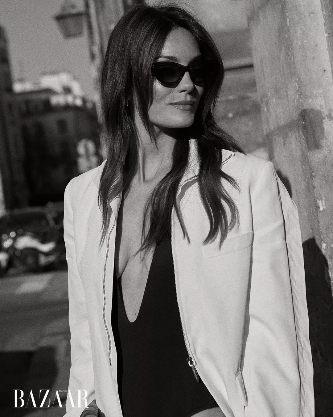 A woman wearing sunglasses and a light-colored jacket over a dark top, leaning against a wall in an urban setting.