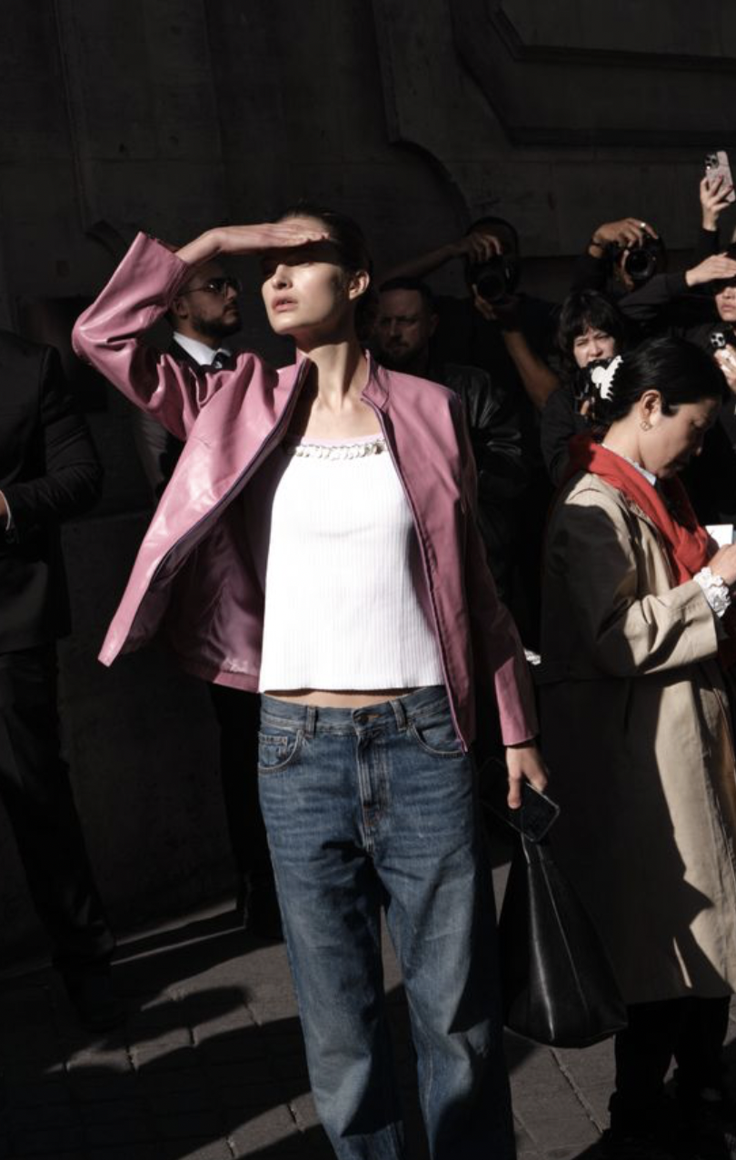 A woman wearing a pink jacket and jeans, shielding her eyes from the sun with her hand in a crowd of people, some holding cameras, in an outdoor setting.
