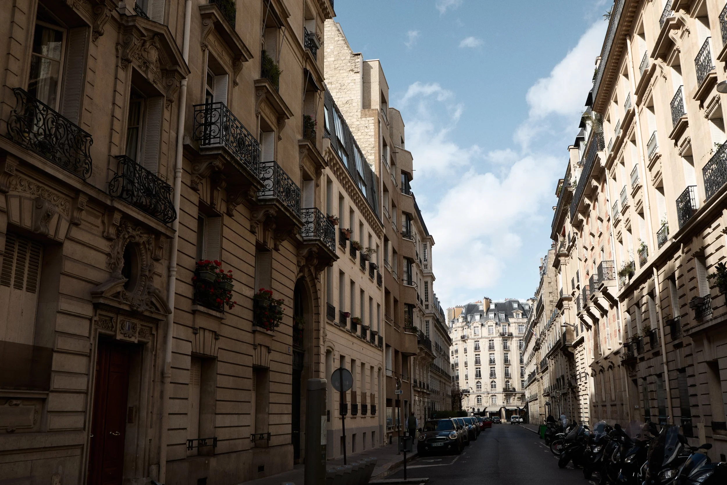 A narrow city street lined with tall, historic, European-style buildings on both sides. Parked cars and scooters are along the street, with a few pedestrians visible. The sky is mostly clear with scattered clouds.