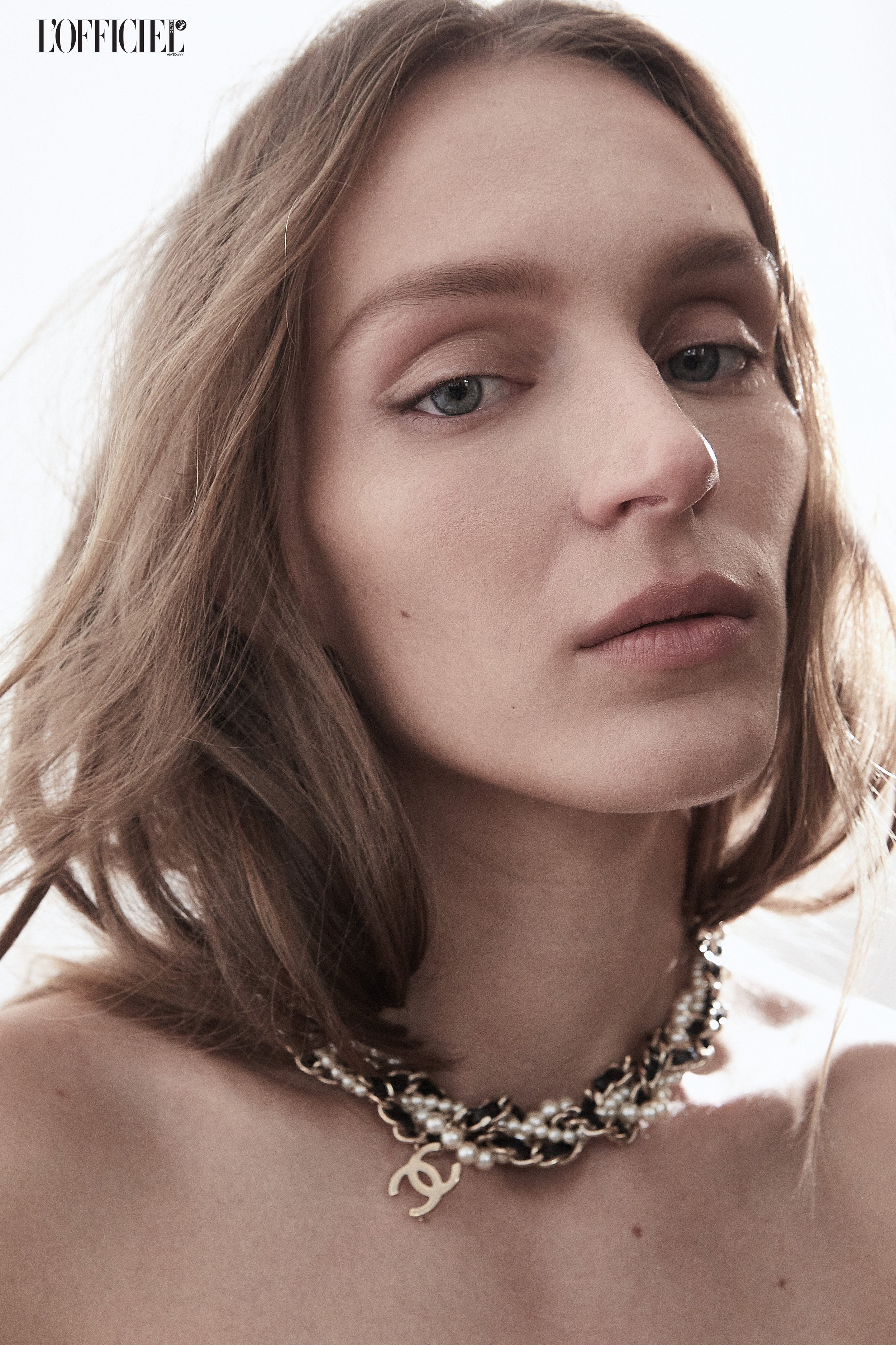 Close-up of a woman with light skin and brown hair wearing a pearl and chain necklace with a Chanel logo pendant, against a neutral background.
