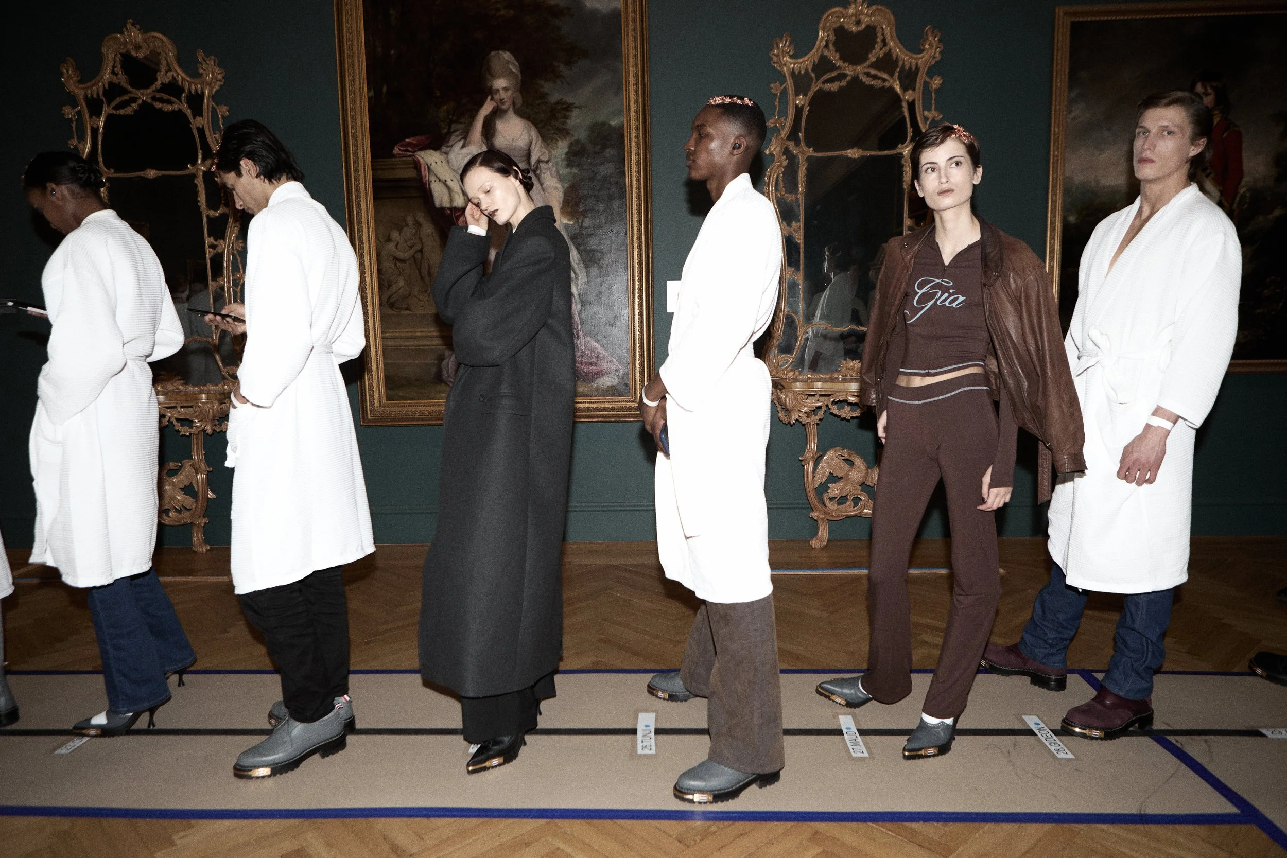A line of mannequins dressed in various clothing styles, displayed in an art gallery with ornate mirrors and framed paintings on the wall behind them.