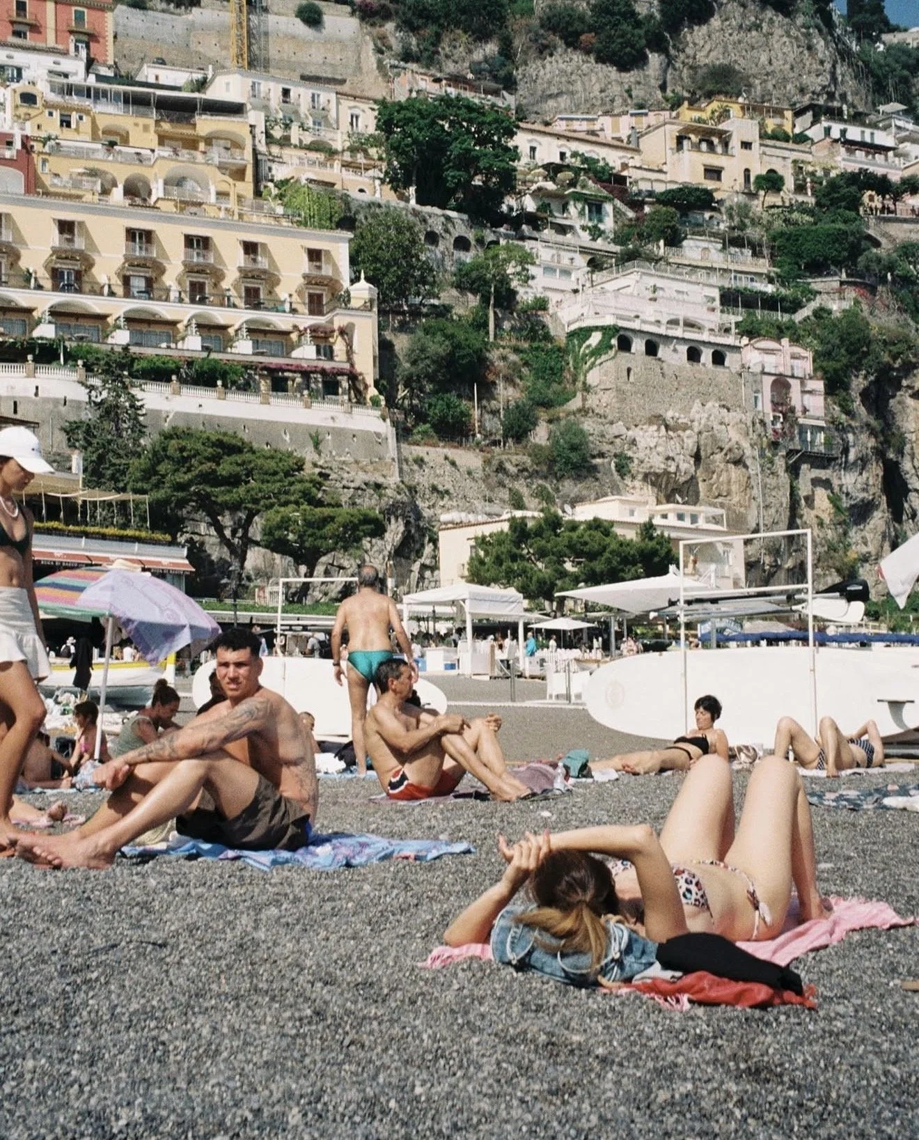 People lounging and sunbathing on a pebbled beach with colorful umbrellas and beach towels, with buildings and lush green trees on a steep hillside in the background.