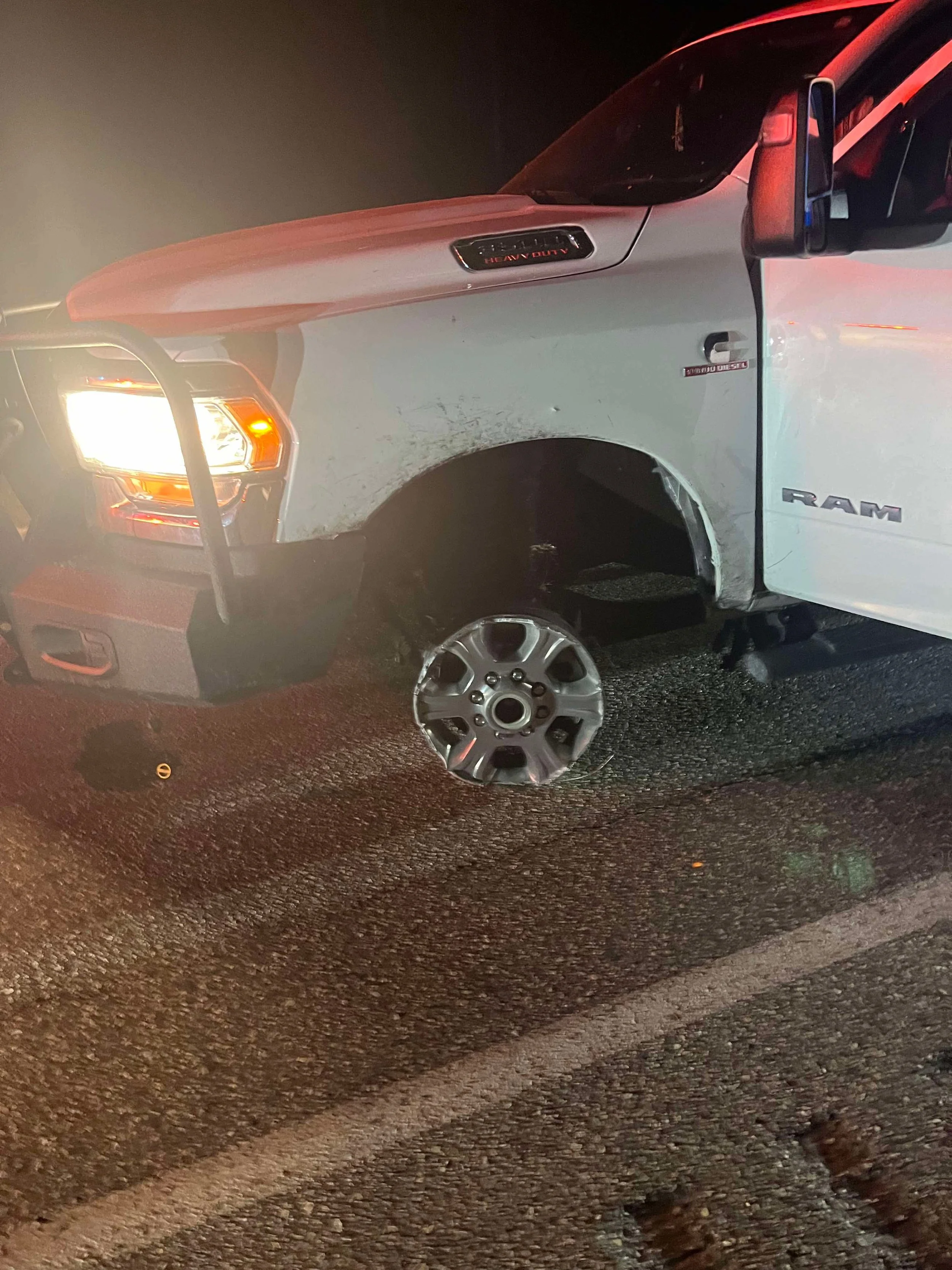 A white RAM pickup truck with damage to the front left wheel area, missing the wheel, with the hub exposed, and the tire on the ground nearby. The truck is on a dark road at night.