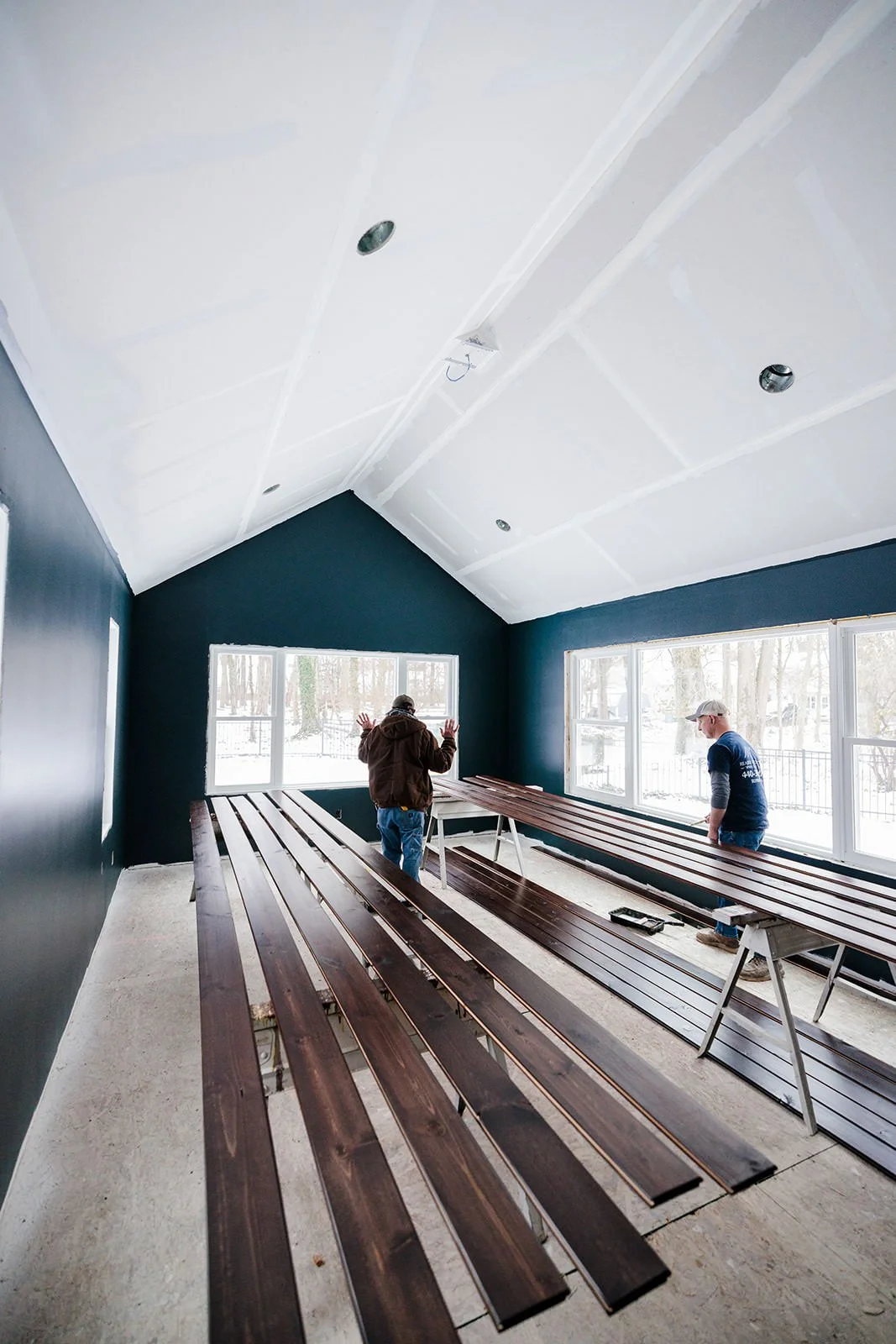 Two men installing dark stained wooden planks on tables in a room with dark blue walls, large windows, and a high ceiling under construction.