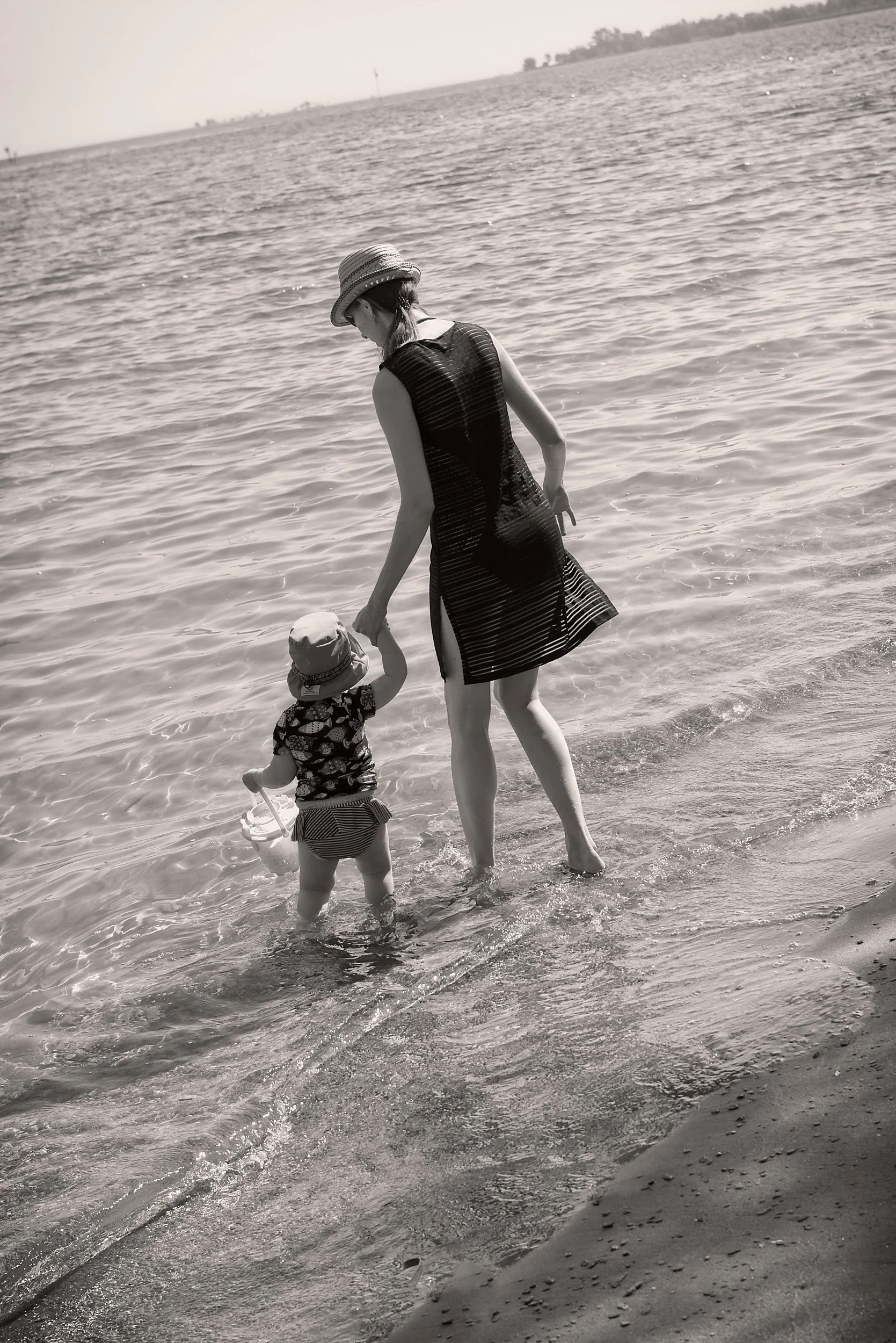 A woman and a small child holding hands while wading in shallow water at the beach, both wearing hats, with the woman in a sleeveless dress and the child in a patterned shirt and shorts.