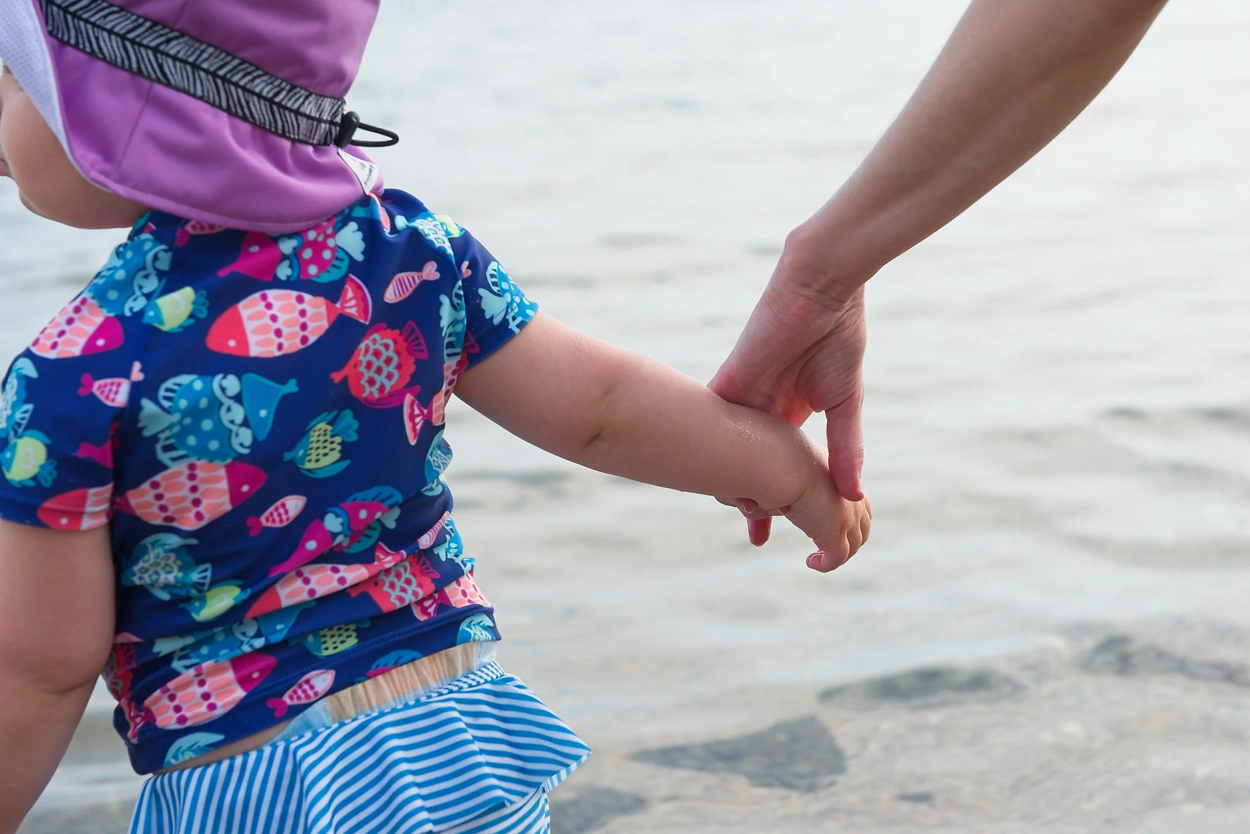 A child in a colorful fish-patterned shirt and striped bottoms holding hands with an adult near the water.
