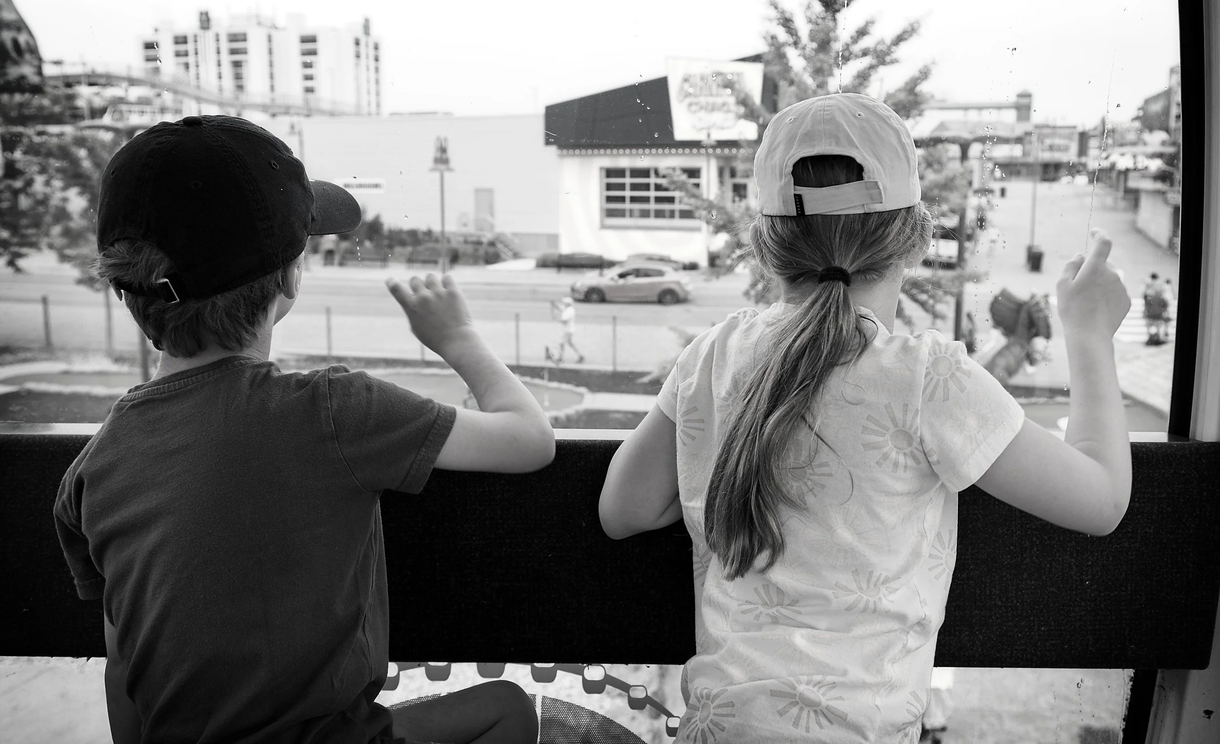 Two children, a boy and a girl, sit at a window looking out at a city street. The boy, wearing a cap and a t-shirt, gestures with his hand. The girl, wearing a cap backwards and a patterned shirt, also points outside.