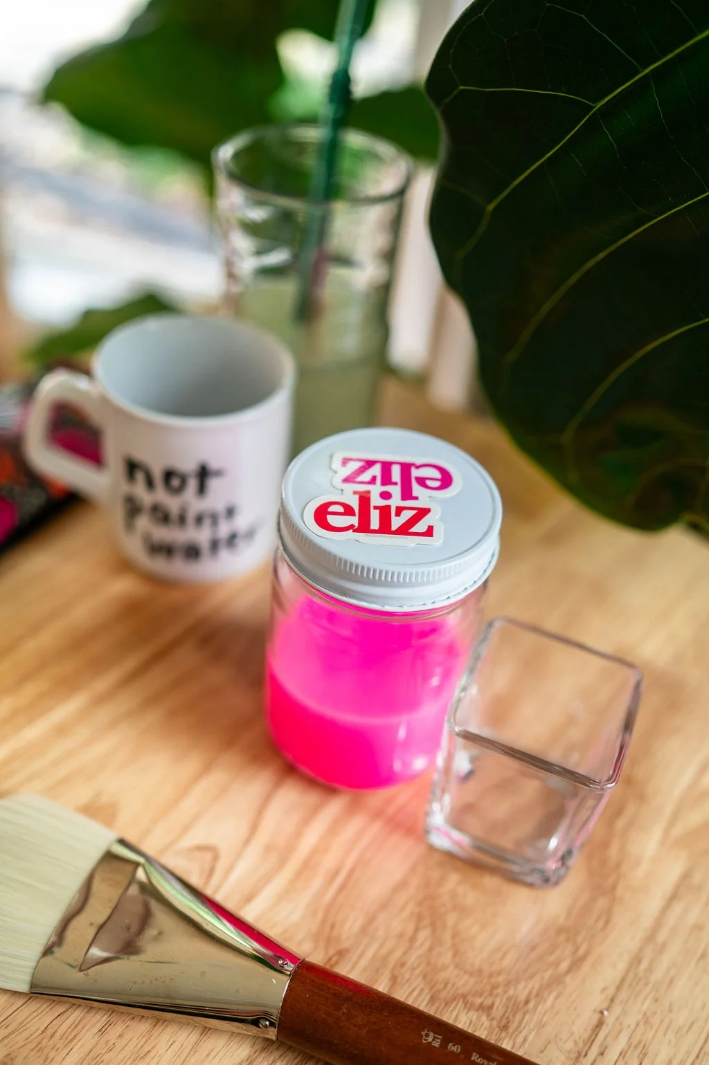 A wooden table with a jar of pink paint, a paintbrush, a white mug with the text 'not paint water', a clear glass, a square glass holder, and plants in the background.