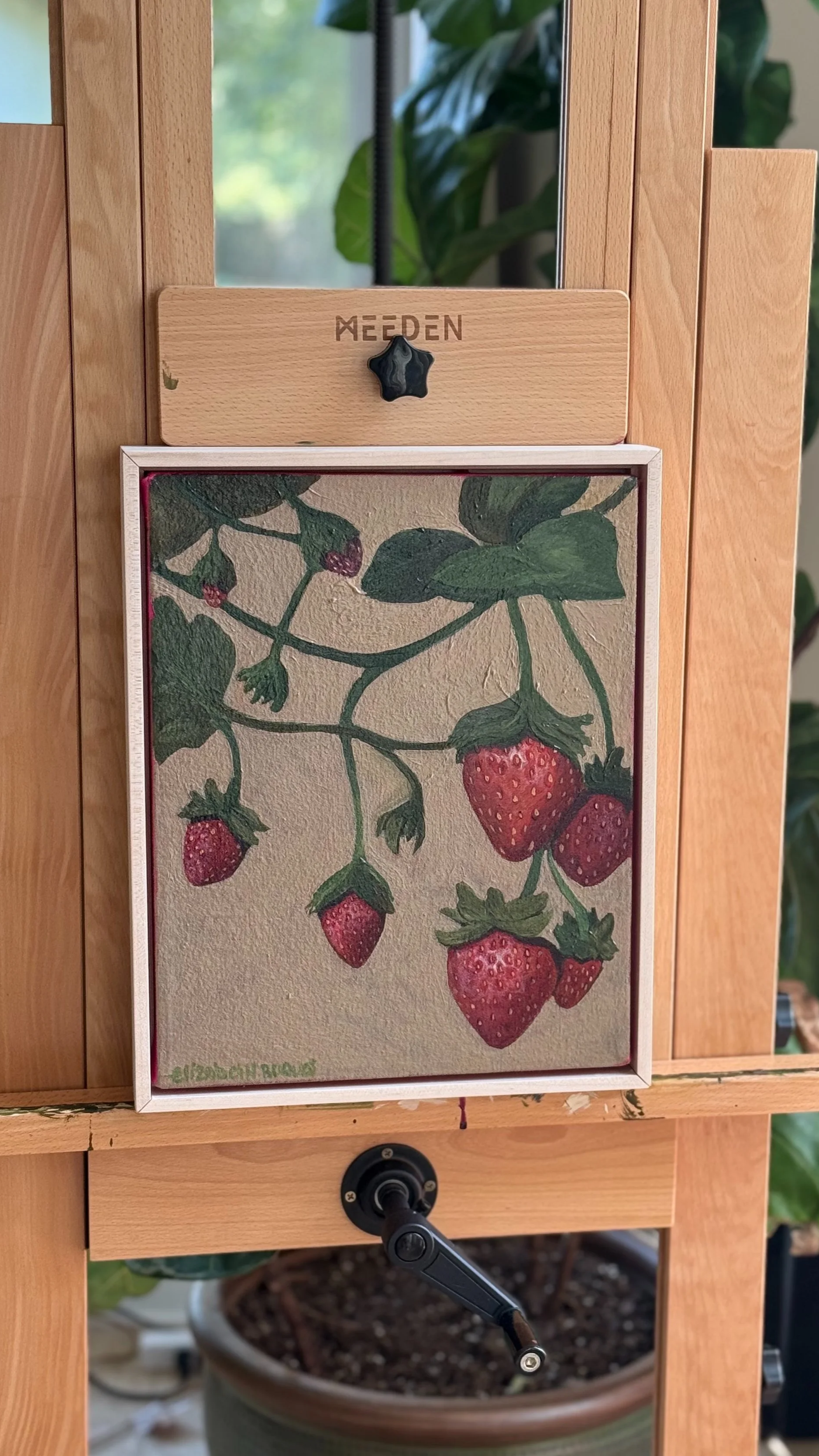 A wooden frame with a painting of strawberries hanging on a light-colored wooden easel. A potted plant with large green leaves is visible in the background.