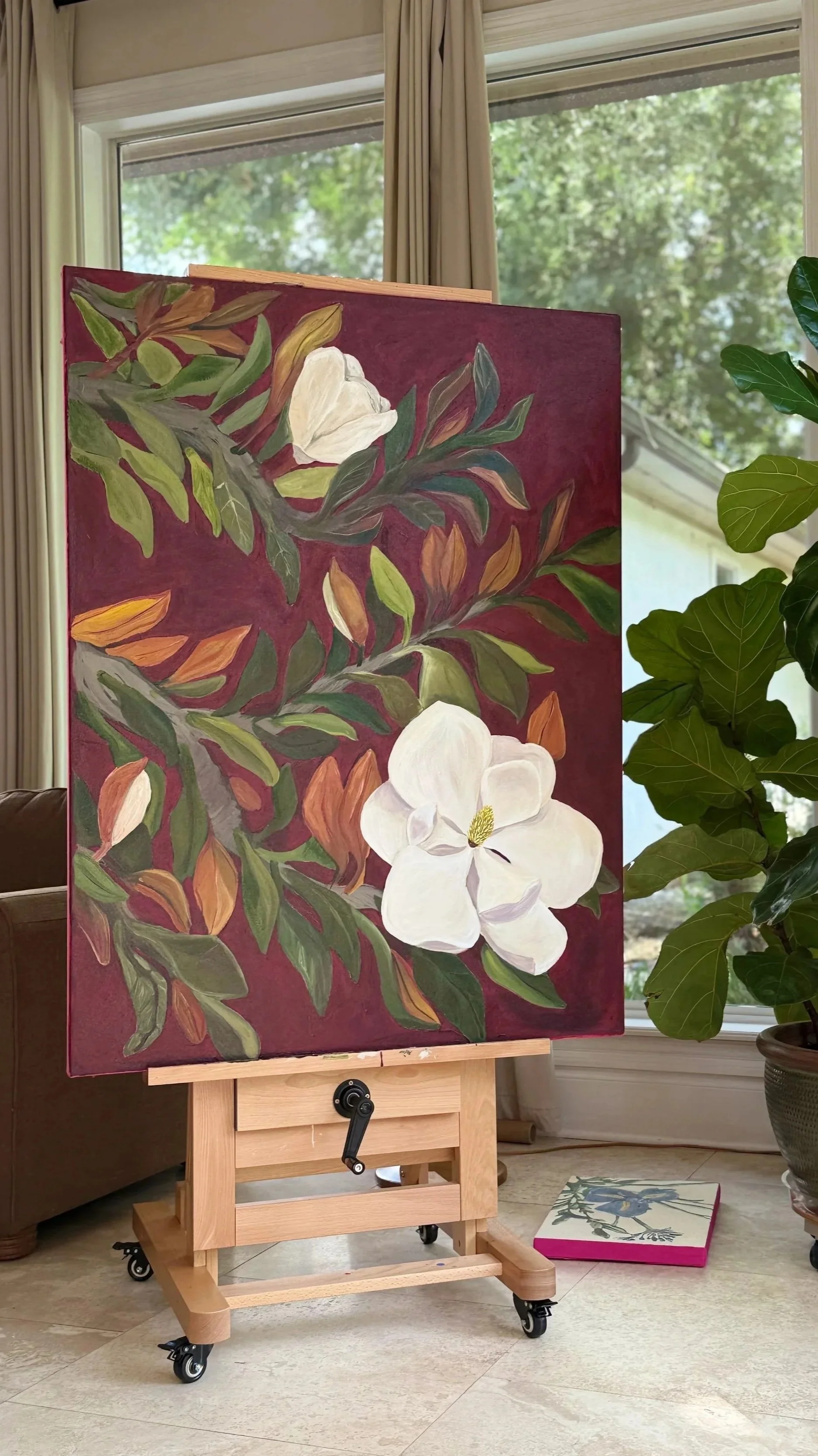 An easel with a painting of white magnolia flowers and green leaves on a maroon background, set indoors near a window with curtains, next to a plant in a pot.