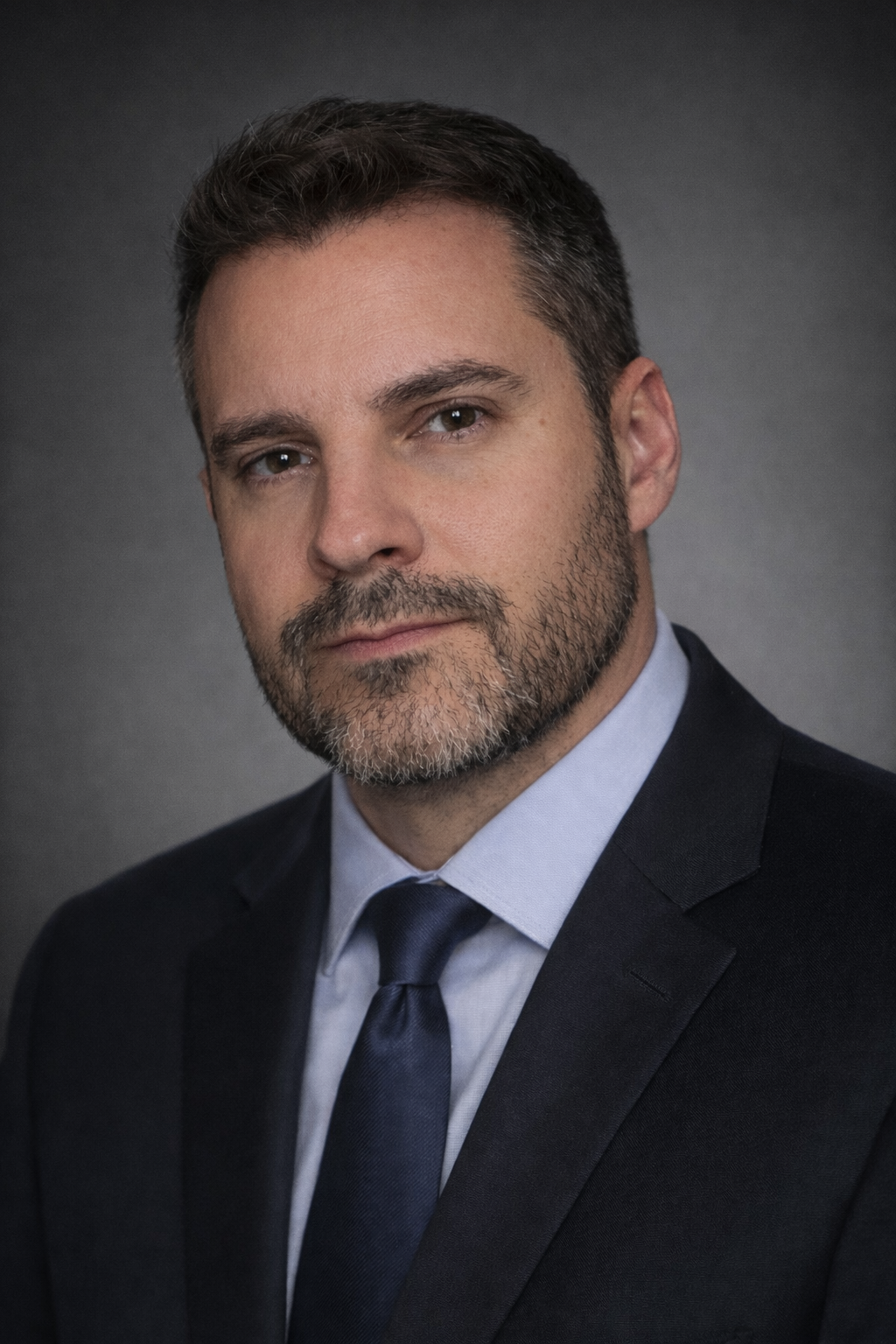 Headshot of a man with short dark hair, beard, wearing a dark suit, light blue shirt, and dark tie, against a gray background.