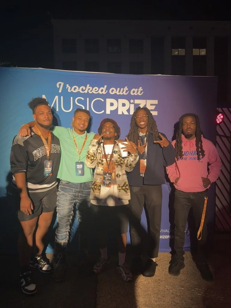 Five young men standing in front of a backdrop at a music award event, smiling and posing for the photo at night.