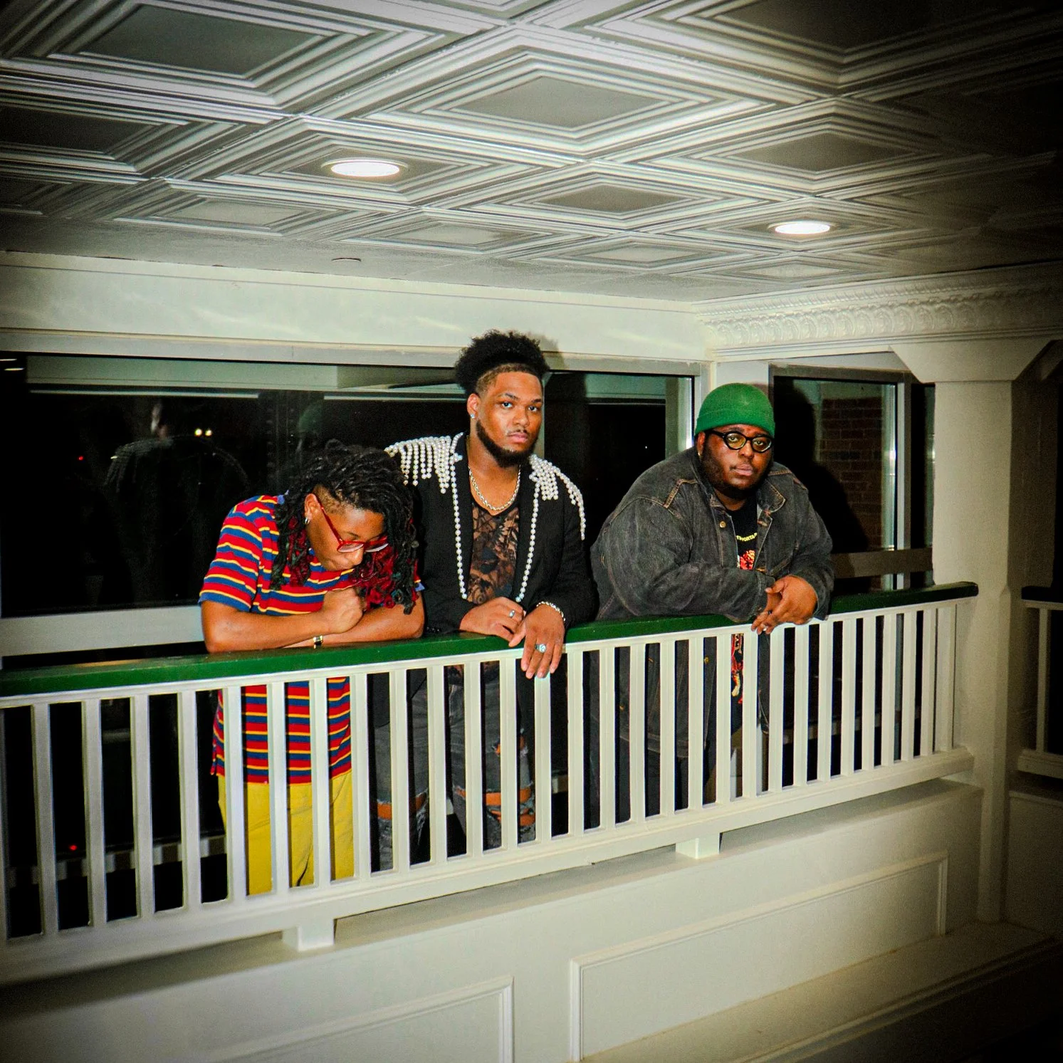 Three individuals standing behind a white railing indoors at night, looking down, with a window behind them showing darkness outside.