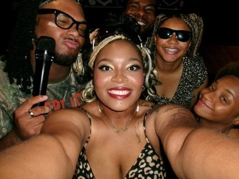 Group of five diverse smiling friends taking a selfie together, with some wearing sunglasses and fashionable clothing.