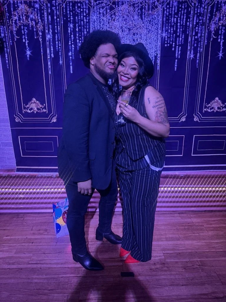 Two people smiling and posing together at an event with purple decorative lighting, one holding a glass of drink, both dressed in stylish clothing. The man has curly hair and is in a black blazer and pants, while the woman has dark hair and tattoos, 