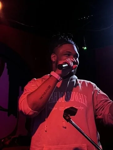 A man wearing a red hoodie speaking into a microphone on stage at night.