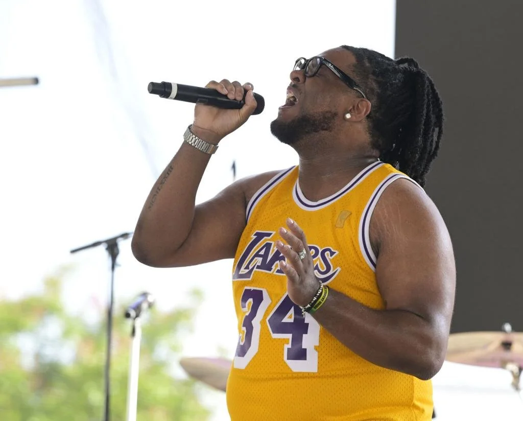 Man singing into a microphone while wearing a Los Angeles Lakers basketball jersey with the number 34, black glasses, and dreadlocks.