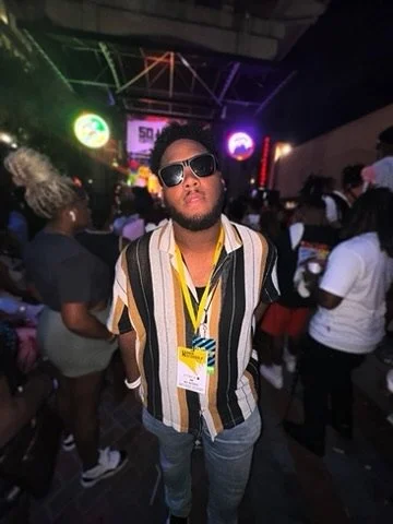 Man wearing sunglasses, striped shirt, and lanyard at an indoor event with colorful neon lights and a crowd.