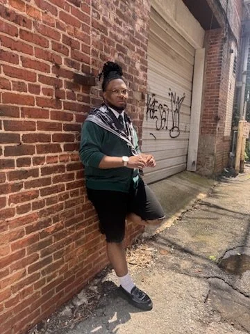 Person standing against a brick wall in an alleyway, wearing glasses, a green and black hoodie, black shorts, white socks, and black sneakers.