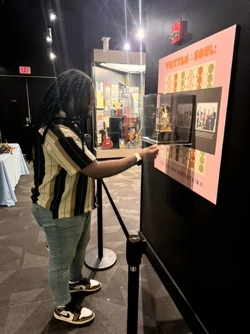 A woman in a black and beige striped shirt and jeans touching a digital touchscreen display at an indoor event or museum, with a sign saying "VOTING: SOUL" on the wall beside her.