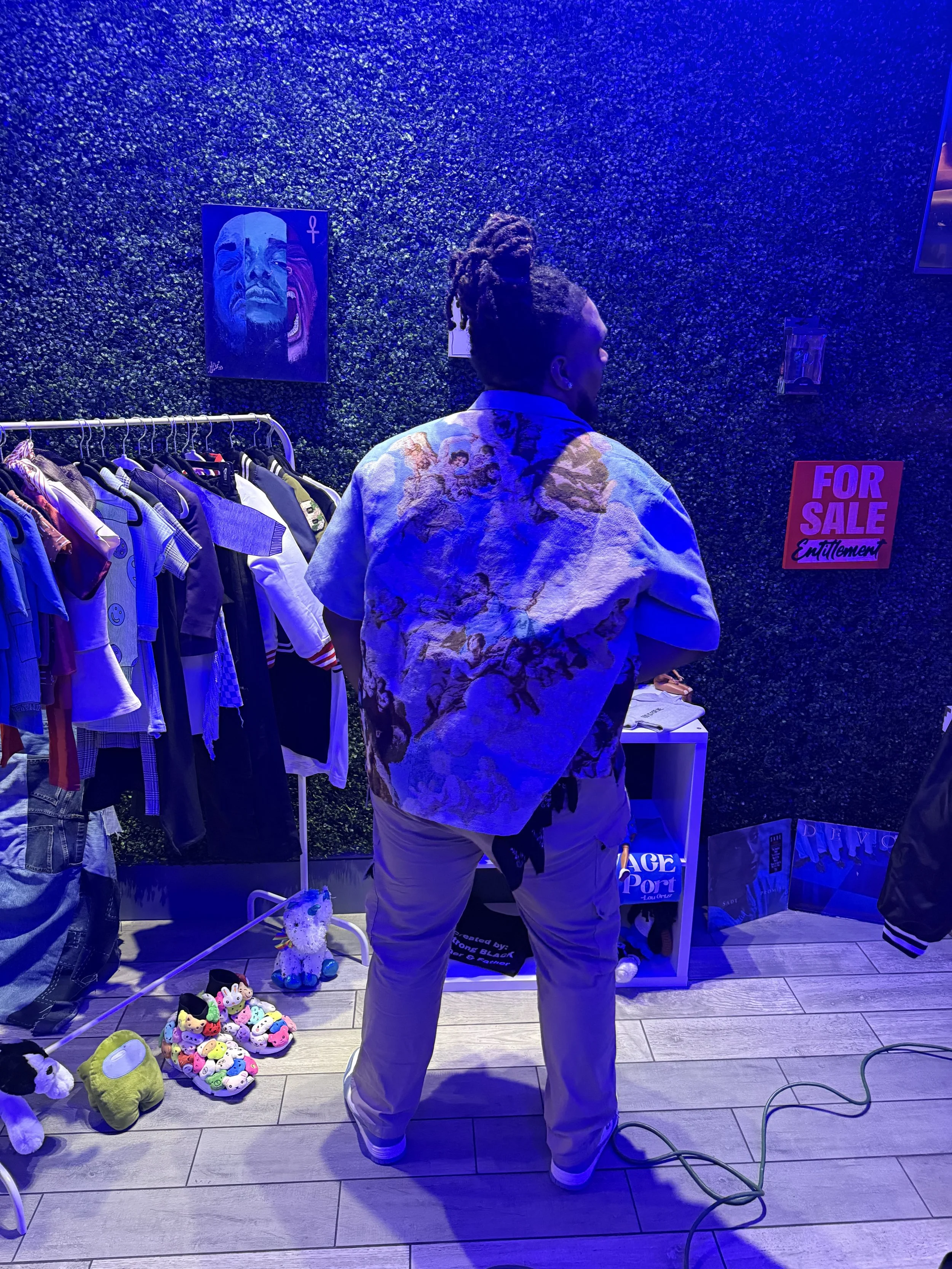 A man with dreadlocks standing in a clothing store amid apparel and plush toys, under blue lighting, with a textured green wall background and a "For Sale" sign.