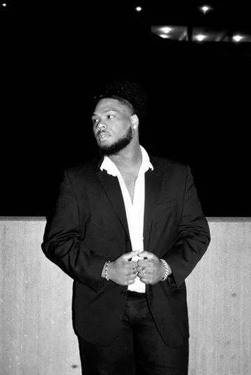 A man in a suit standing against a dark background, looking to the side with hands clasped in front.