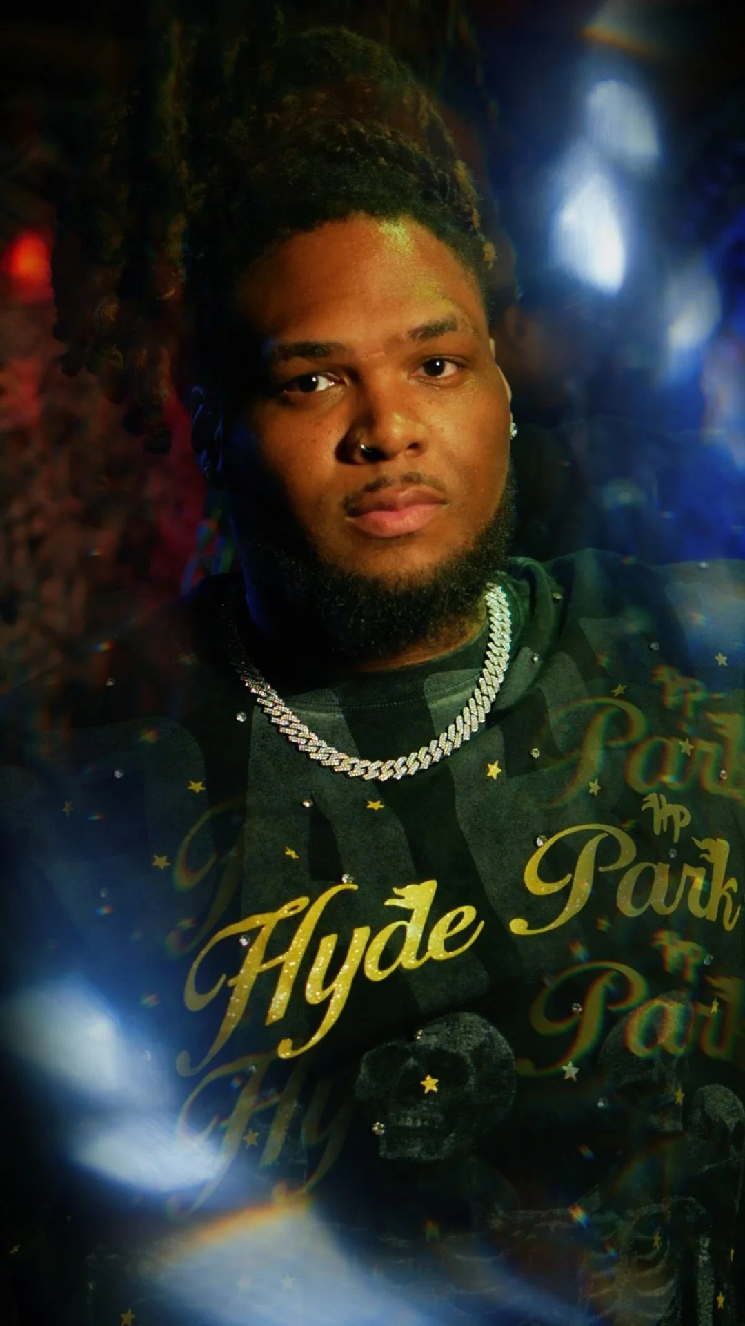 A young man with braided dreadlocks, a nose piercing, and a beard takes a selfie in a dimly lit setting. He's wearing a dark shirt with yellow text that reads 'Hyde Park' and a thick, shiny chain necklace.