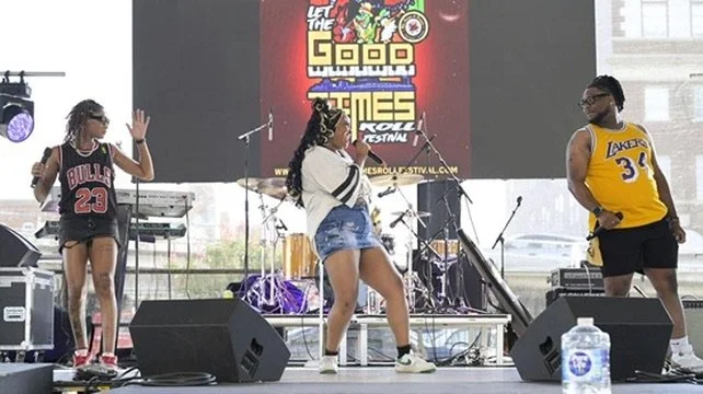 Three young performers on stage during a live music event, with musical equipment and a large screen displaying event graphics in the background.