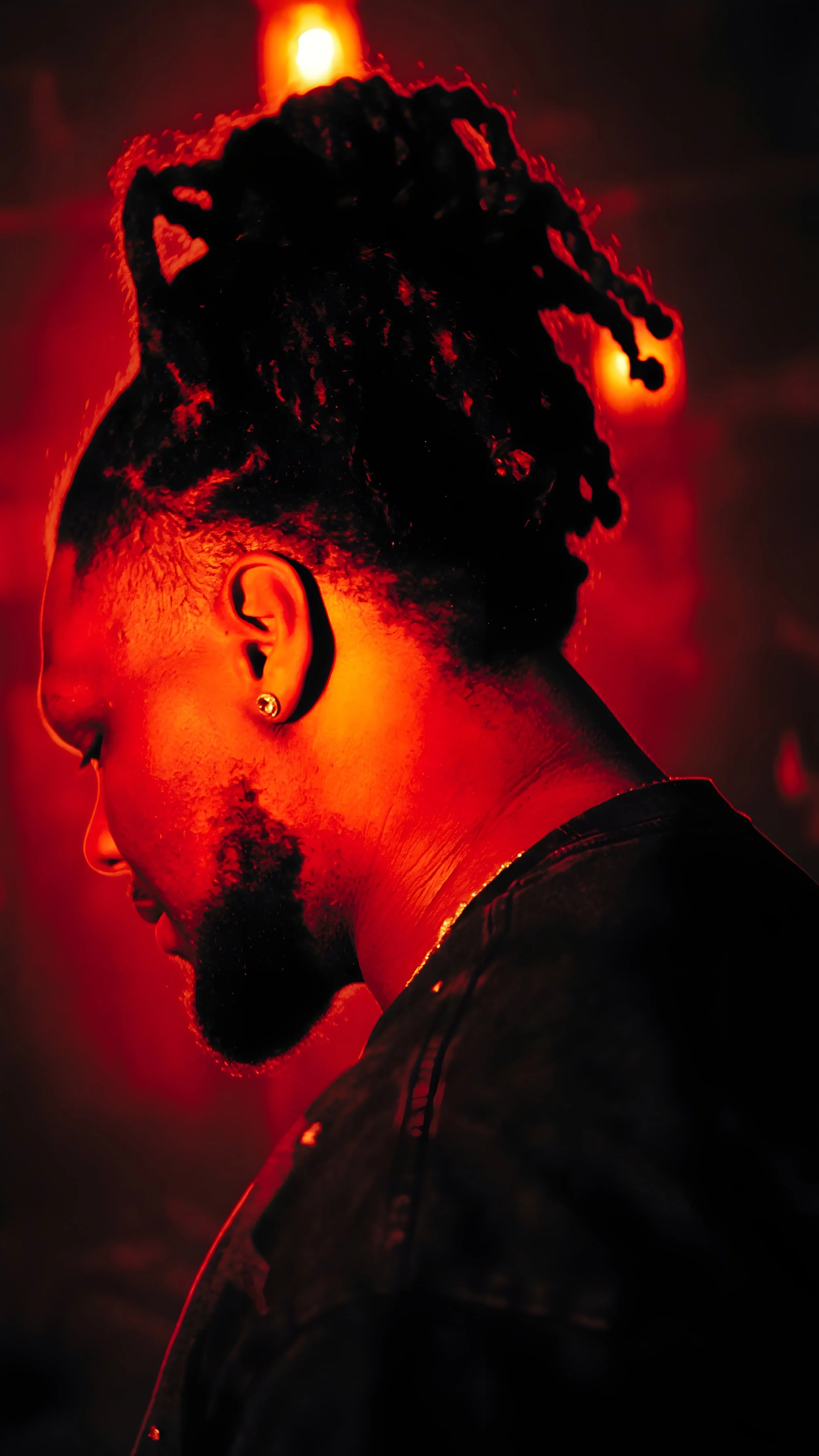 Profile of a man with dreadlocks, earring, and facial hair looking down in a red-lit environment.