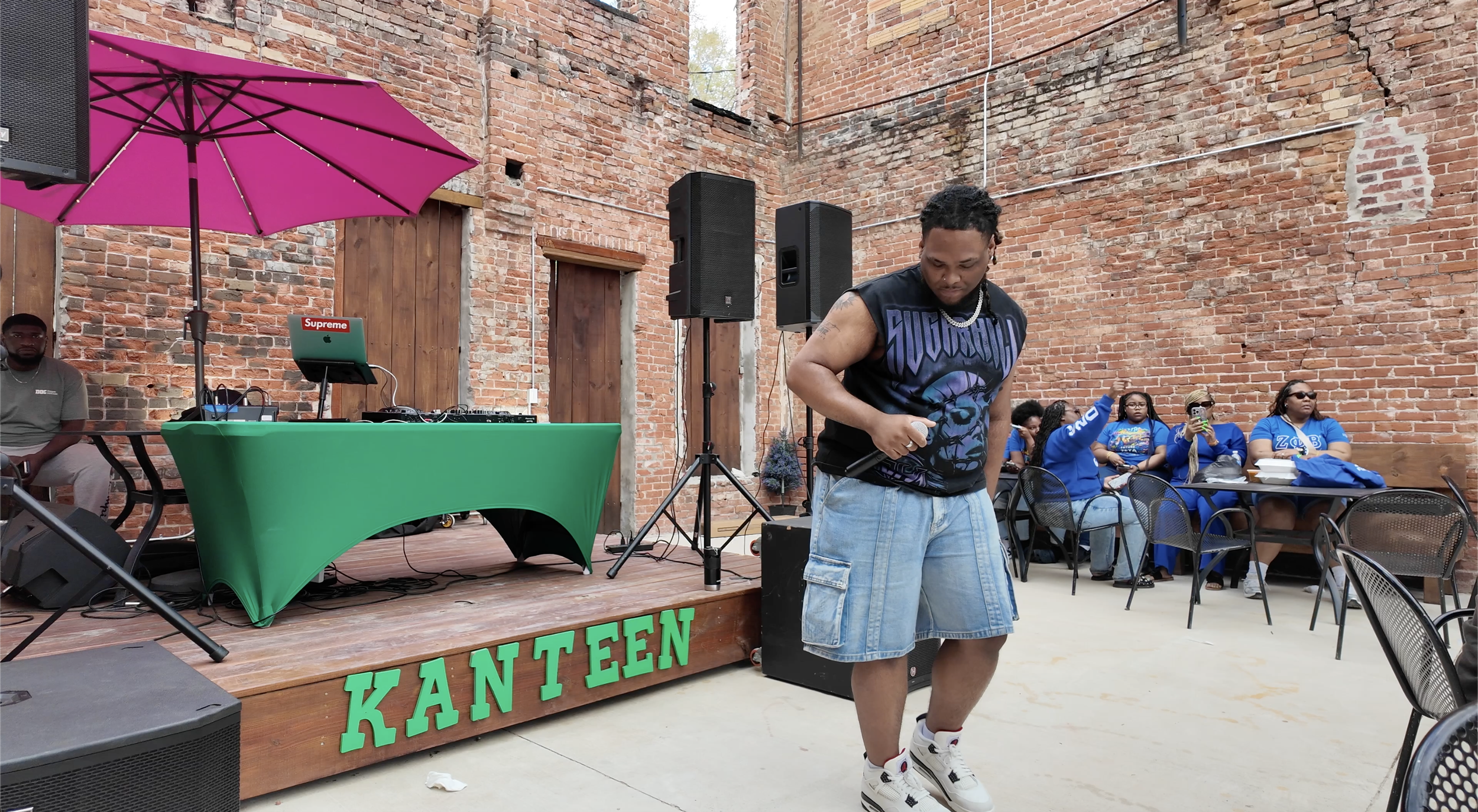 Man in sleeveless shirt and shorts holding microphone, standing on small stage with green 'KANTEN' sign, set up with DJ equipment and speakers, brick wall background, audience seated nearby, some taking photos.