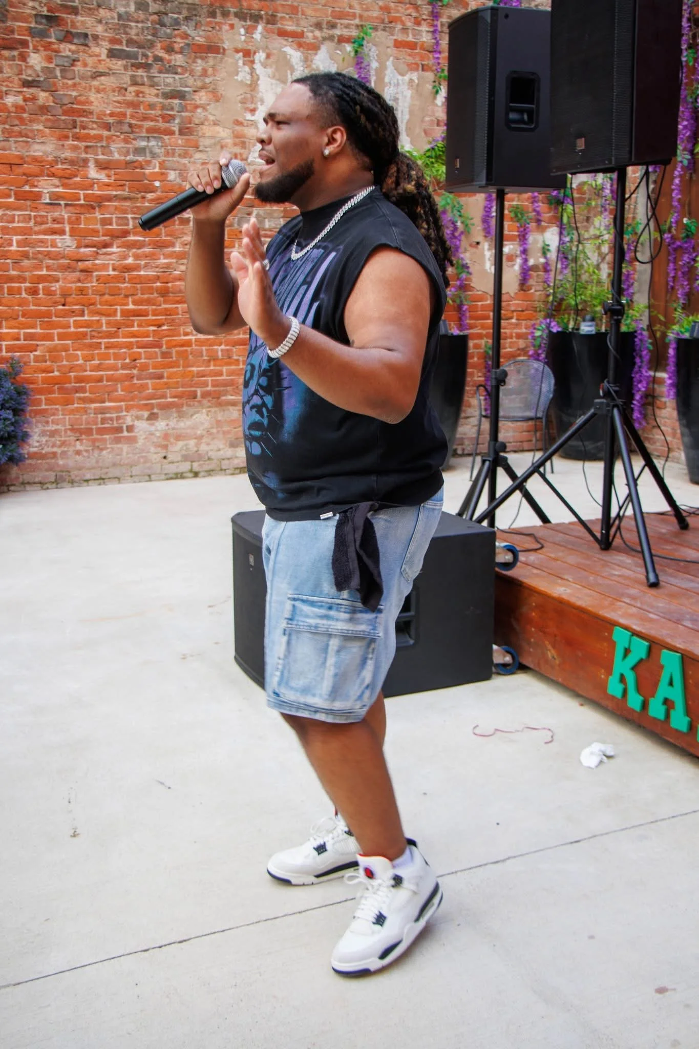 A man singing into a microphone during an outdoor performance. He is wearing a sleeveless t-shirt, shorts, and sneakers, with large speakers and purple flowering plants in the background.