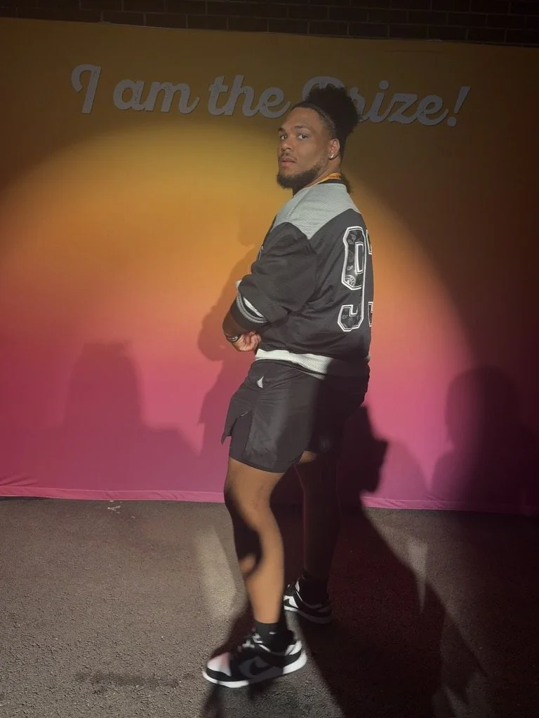 Young man with a beard and tied-up hair, wearing a varsity jacket, shorts, and sneakers, standing against a colorful gradient background with text that says "I am the Guize!"